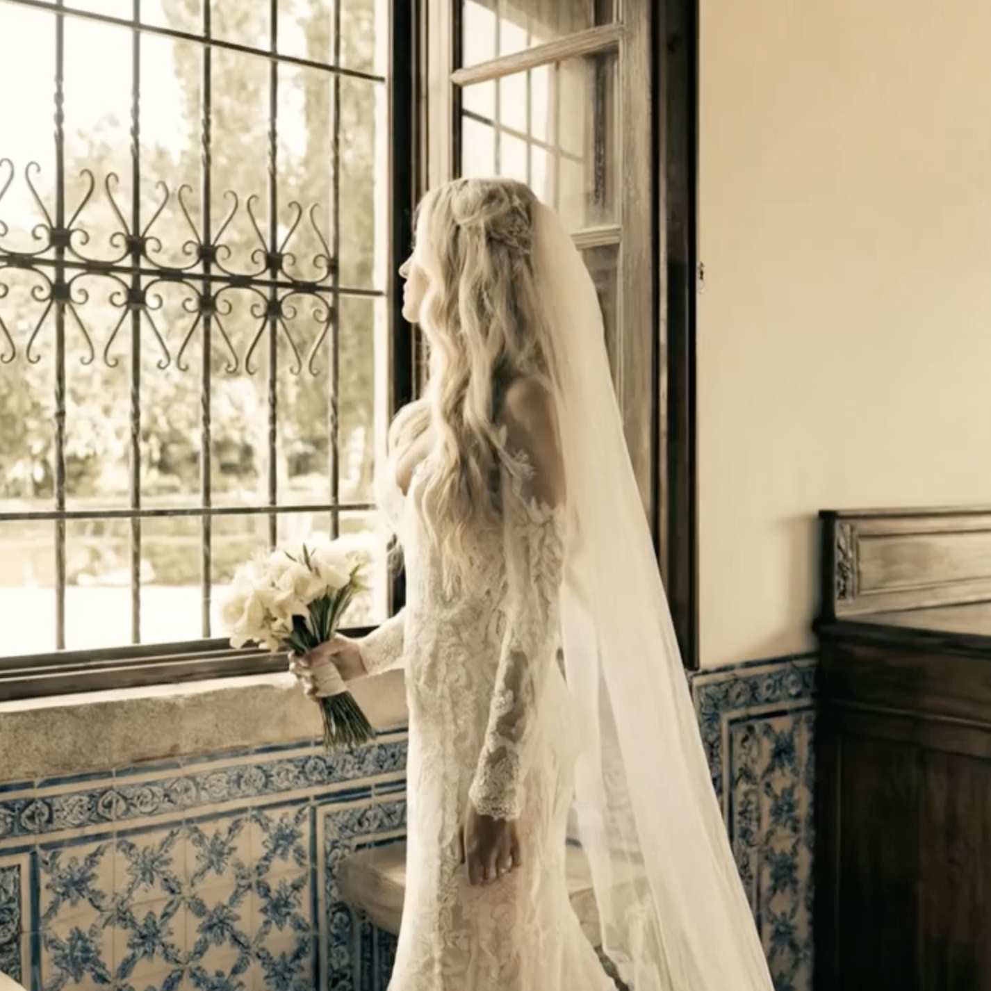 A bride in a lace wedding dress holding a bouquet, standing by a window with decorative ironwork, looking outside on a sunny day.