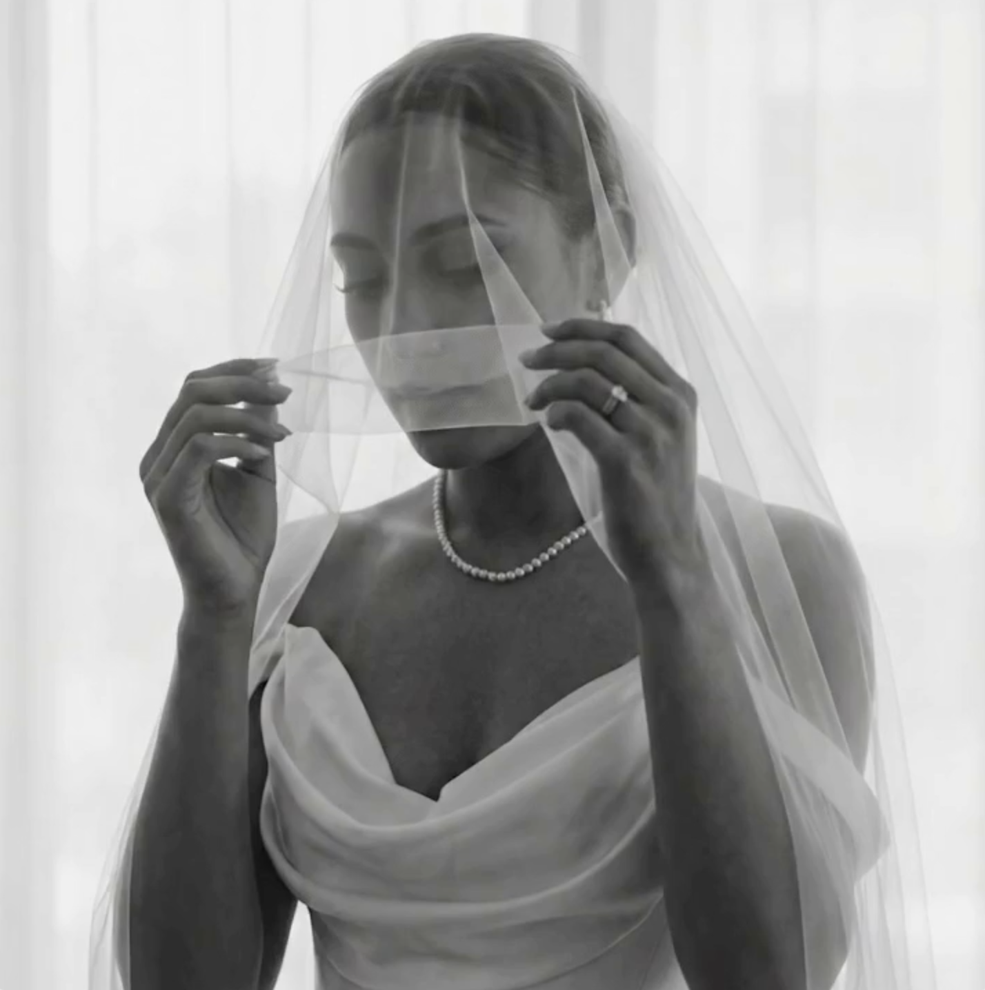 A woman with her eyes closed, wearing a wedding dress, pearl necklace, and a veil, holding the veil with both hands.