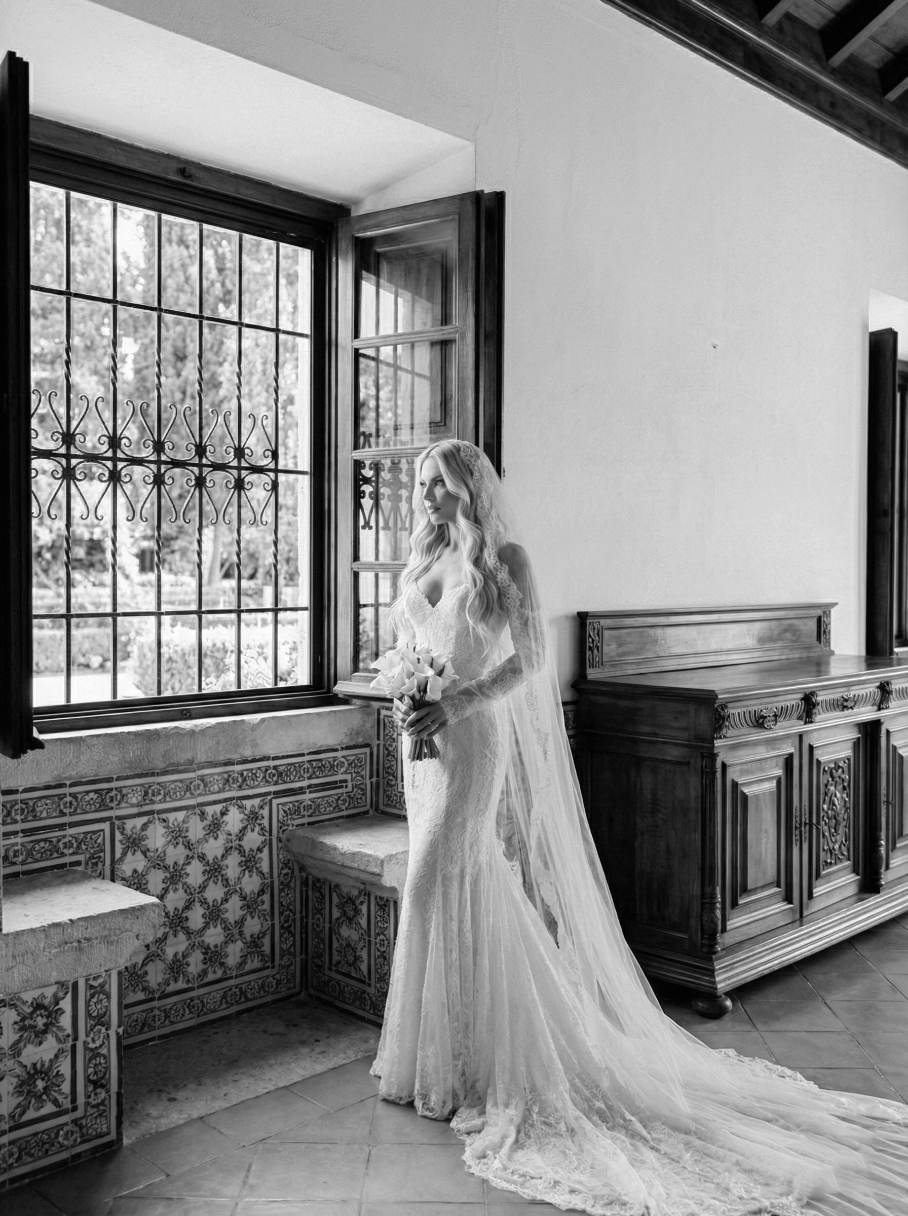 A bride in a lace wedding dress holding a bouquet stands by a window with decorative ironwork, inside a room with ornate wood furnishings and tiled walls.
