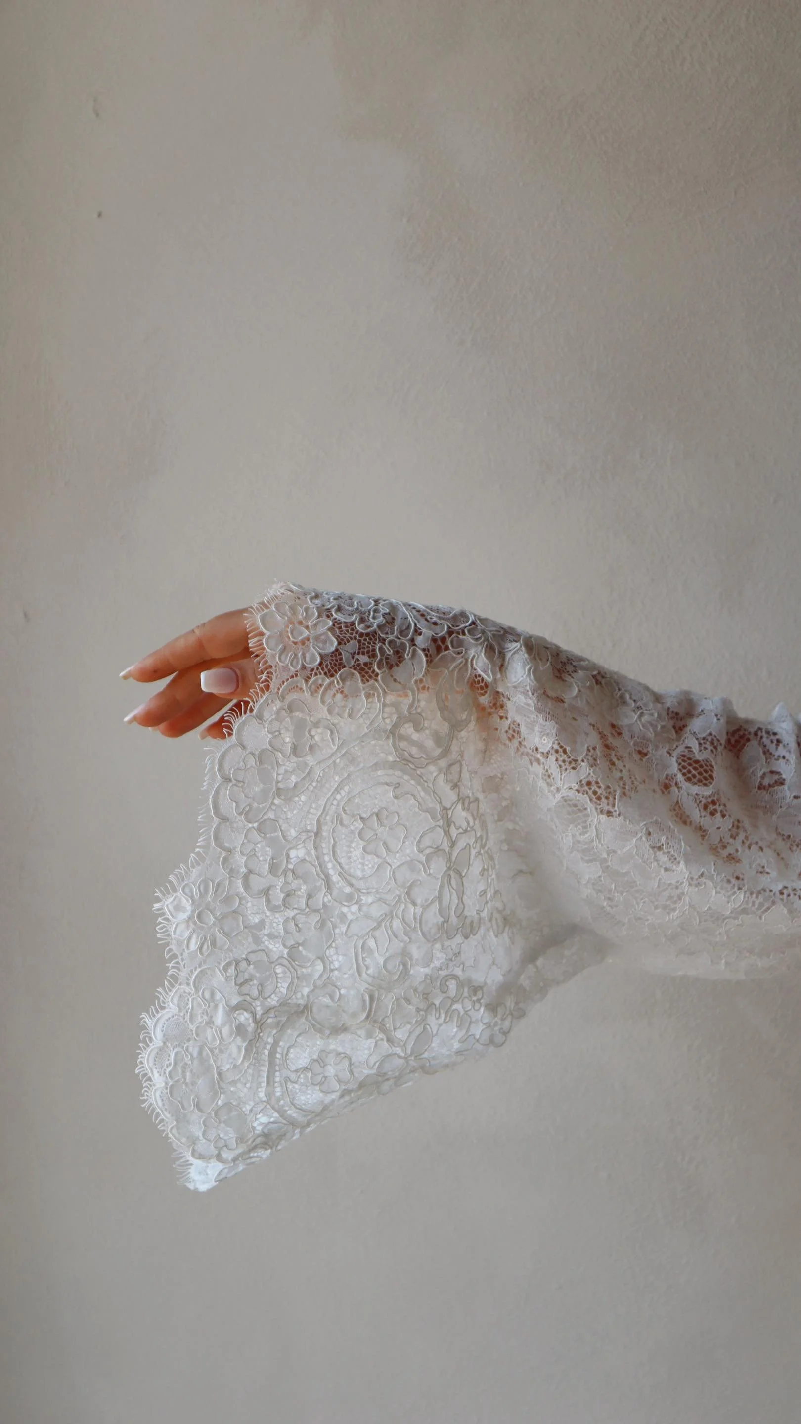 Close-up of a hand with manicured nails wearing a white lace glove or sleeve, against a plain wall background.