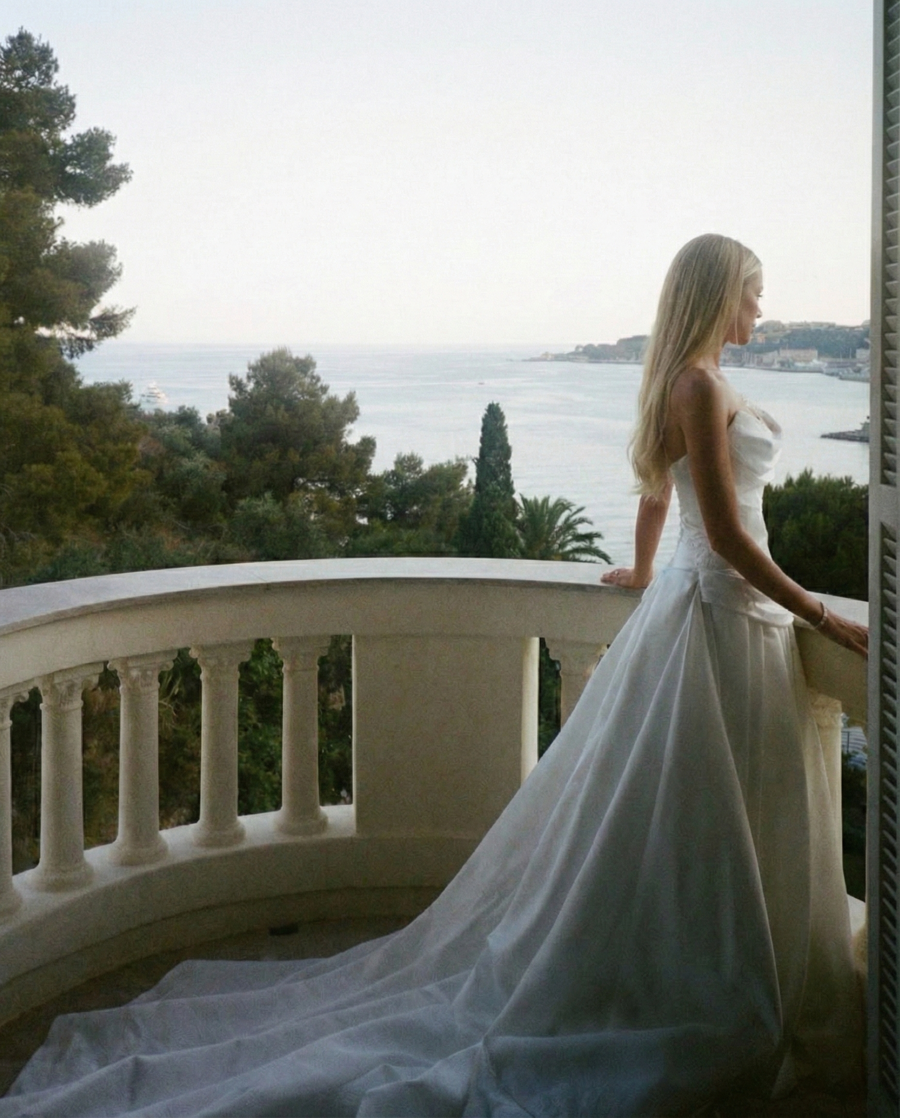 A woman in a white wedding dress standing on a balcony, looking out over a body of water with trees and buildings in the distance.