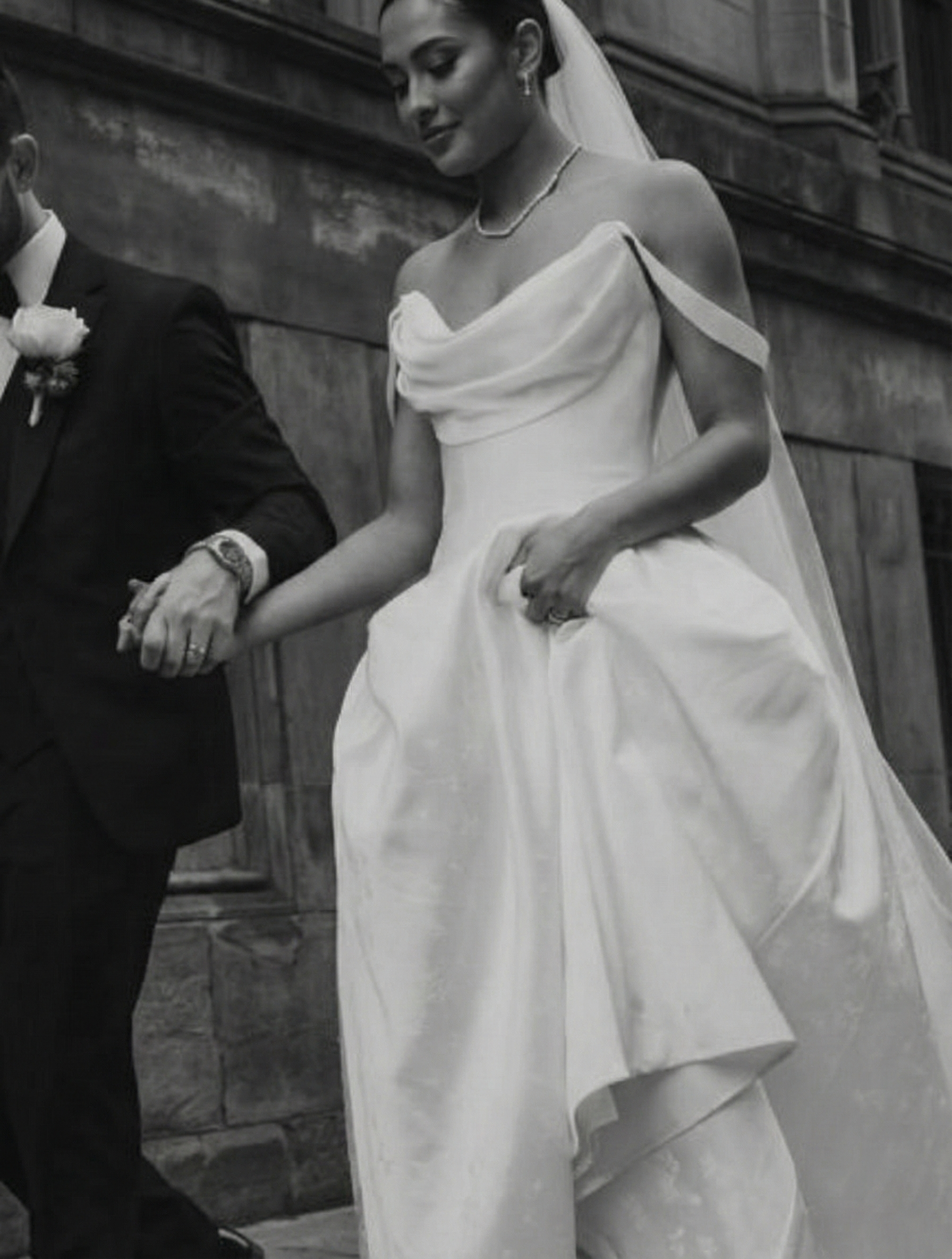 Black and white photo of a bride and groom holding hands during a wedding ceremony. The bride is in a wedding dress, and the groom is in a suit. They are standing outdoors against a stone wall.