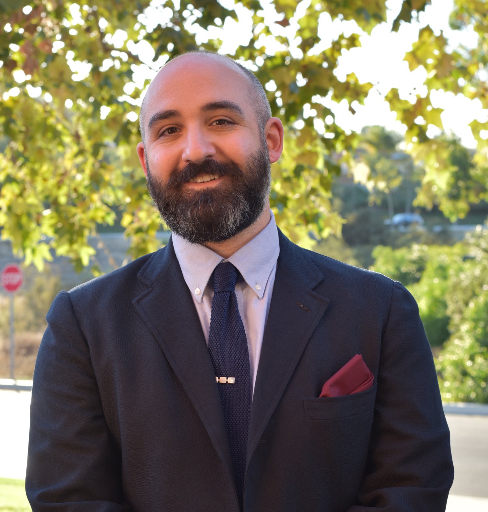 A man with a beard and mustache in a navy suit, white shirt, navy tie, and red pocket square is standing outdoors in front of green trees with sunlight filtering through.