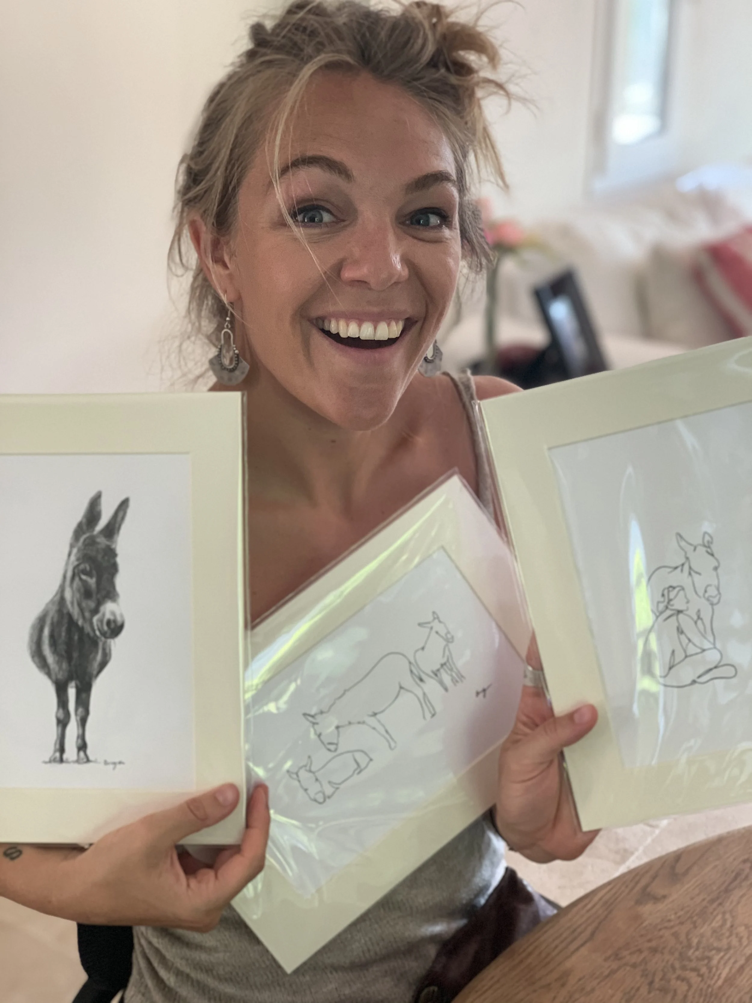A woman smiling and holding up three framed drawings of animals, including a donkey, a goat, and a dog, in a room with a blurred background.