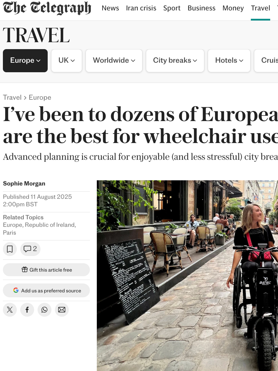 A woman in a wheelchair riding along a cobblestone street with outdoor cafes and restaurants on one side, and a green metal fence on the other. The woman has long blonde hair, is smiling, and is looking upward.