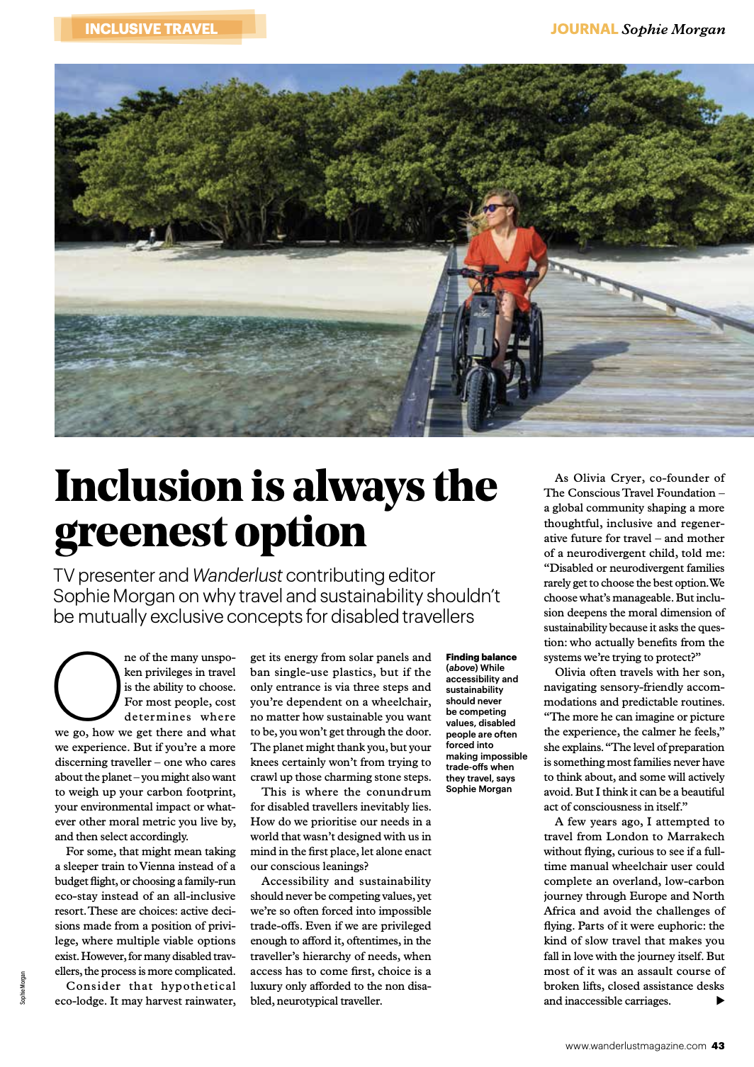 Woman in a red dress in a wheelchair on a wooden pier leading to a beach with trees in the background.