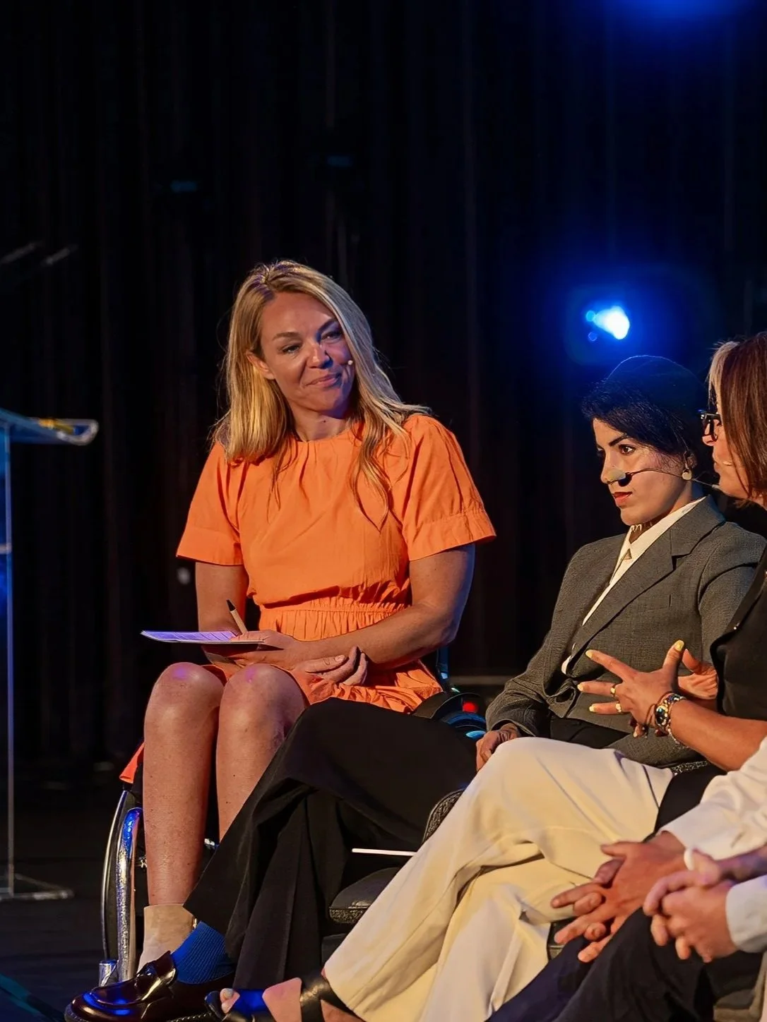 A woman in a wheelchair wearing an orange dress listening to another woman speaking during a panel discussion.