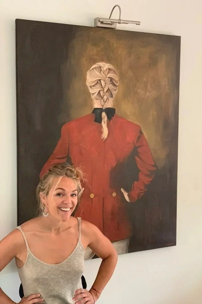 A blonde woman in a beige tank top smiling and posing in front of a large painting of a woman with braided hair wearing a red blazer seen from behind