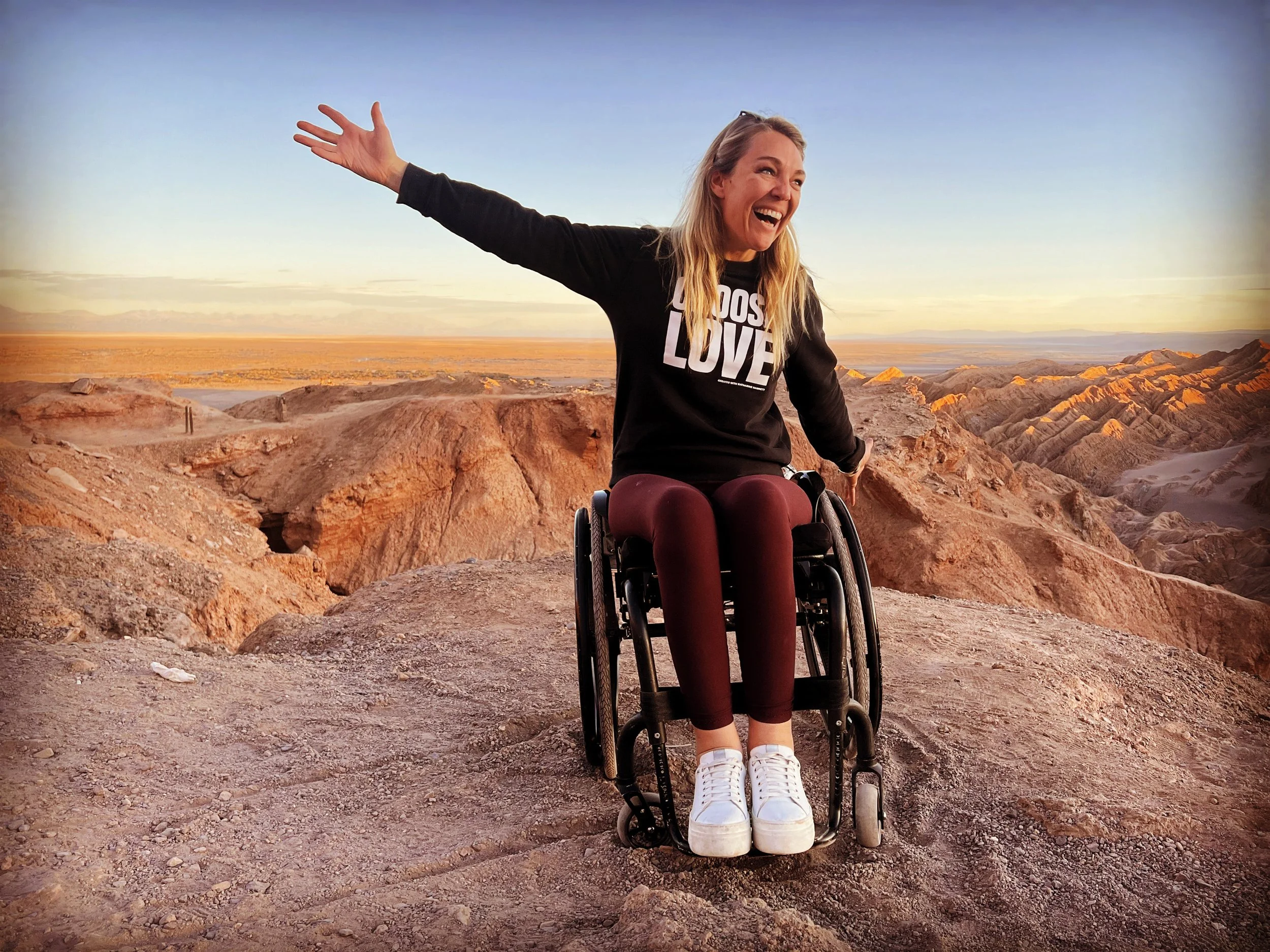 Woman in a wheelchair with outstretched arms smiling, on rocky terrain with mountain ranges and desert landscape in the background during sunset.