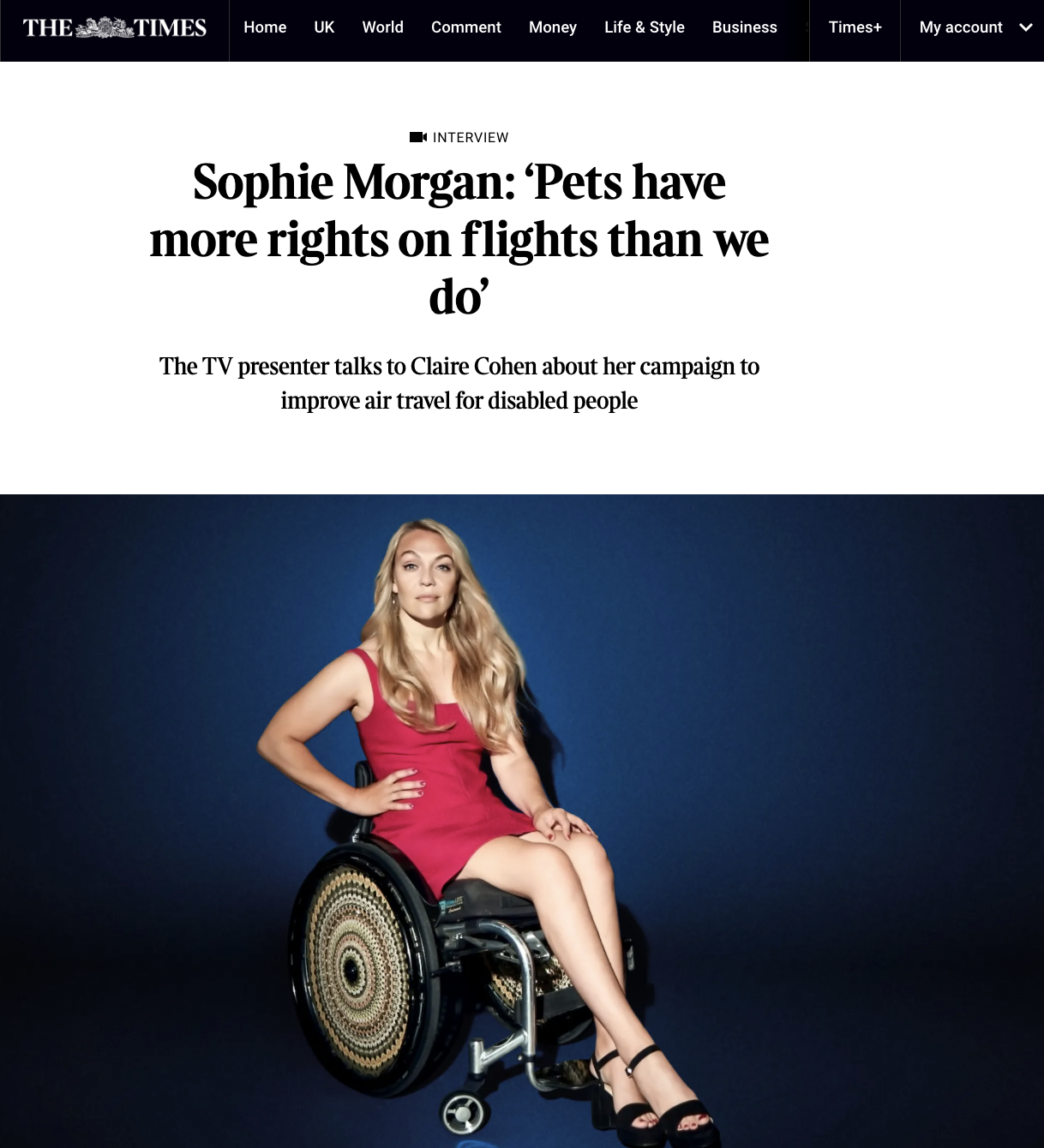 A woman with long, wavy blonde hair wearing a pink dress and black high heels seated in a wheelchair with a decorative wheel design against a dark blue background.