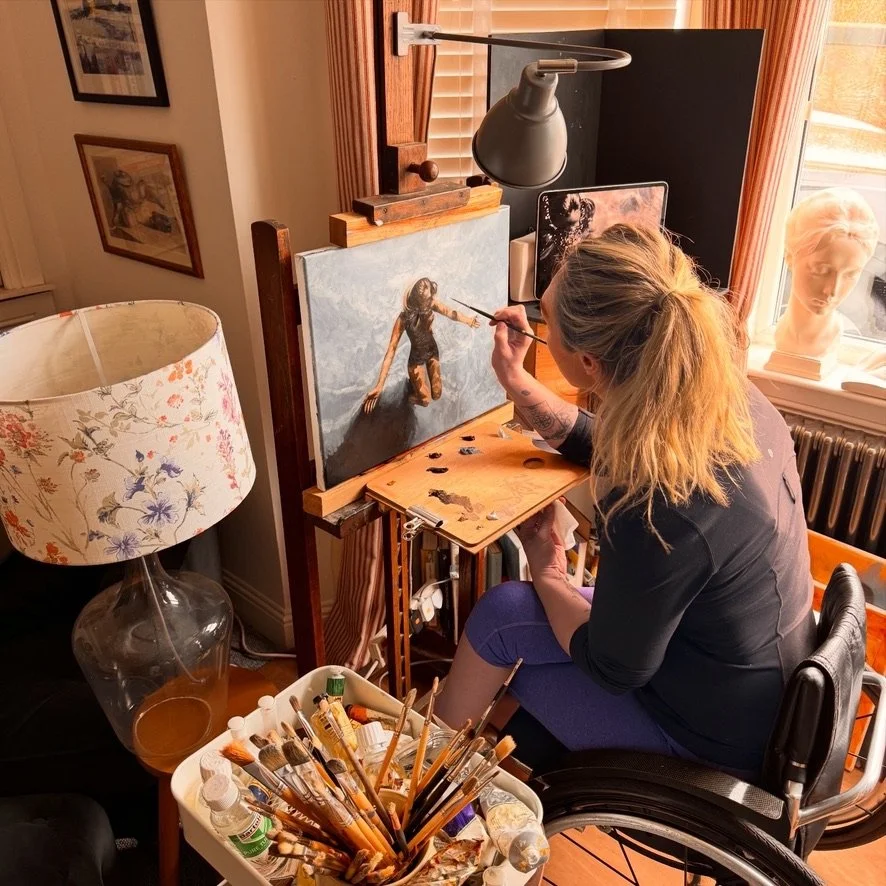 A woman painting a portrait in an art studio, sitting in a wheelchair, with painting supplies and a lamp nearby.