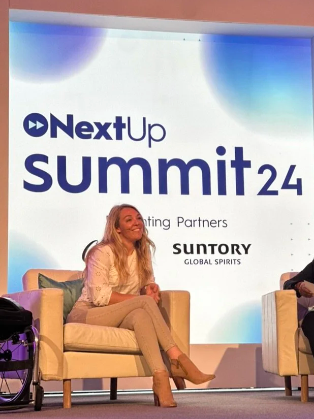 A woman with long blonde hair, wearing a light-colored blouse, beige pants, and brown heels, is sitting on a beige armchair on a stage. She is smiling and looking towards the audience. Behind her, there is a large screen displaying the text 'NextUp summit 24' with sponsor logos, including Suntory Global Spirits.
