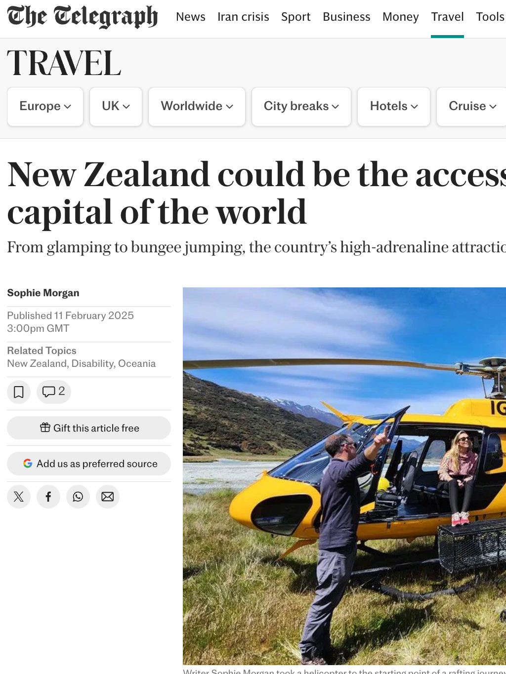 A yellow helicopter with the registration IGY parked in a grassy field with mountains in the background. There is a woman sitting at the open door of the helicopter and a man standing outside, engaging with her. A wheelchair is placed on the ground near the helicopter.