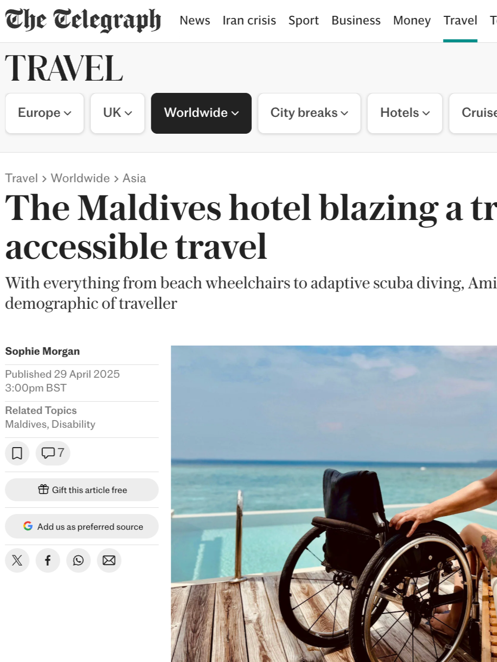 A woman with long hair, tattoos, and sunglasses sitting on a bed by the pool at a beachside hotel, with a disabled beach wheelchair nearby, overlooking the ocean.