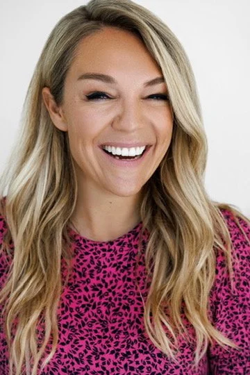 A woman smiling and winking, wearing a pink and black patterned top against a plain background.