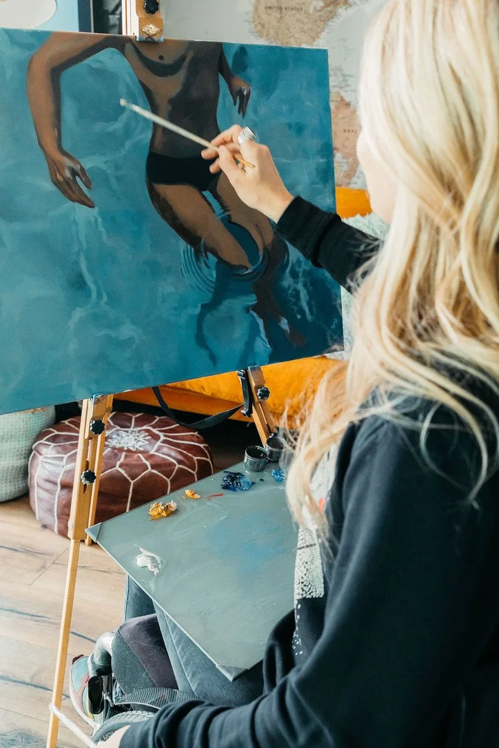 A woman painting a portrait of a person wearing black shorts and a dark top, reflected in water.