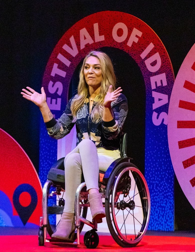 Woman in a wheelchair speaking at the Elevate Festival of Ideas, with colorful event signage in the background.