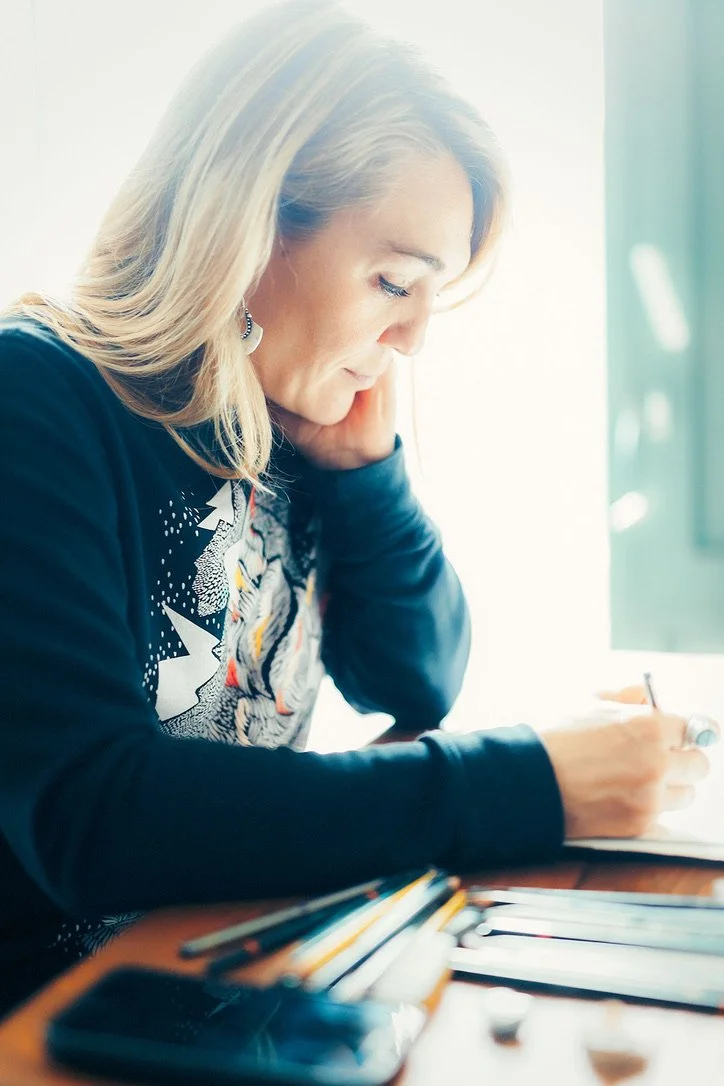 A woman with blonde hair sitting at a table, writing in a notebook, with a smartphone and various pens nearby, in a bright room.
