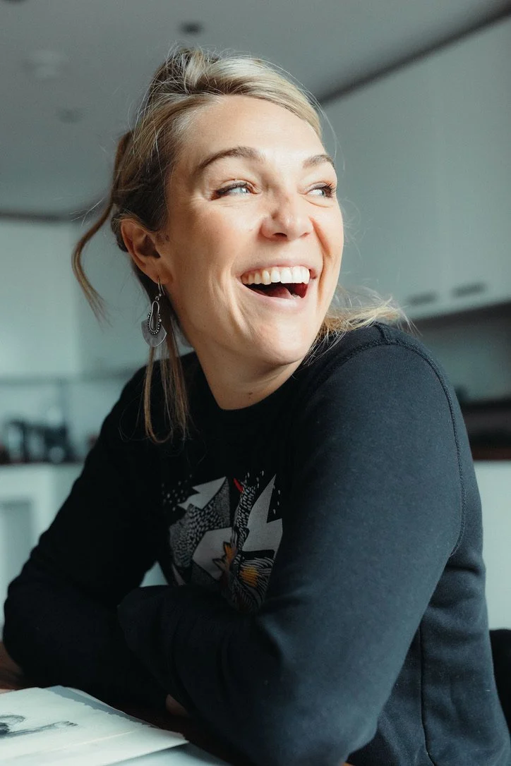 A woman with blonde hair smiling and laughing while sitting at a table in an indoor setting.