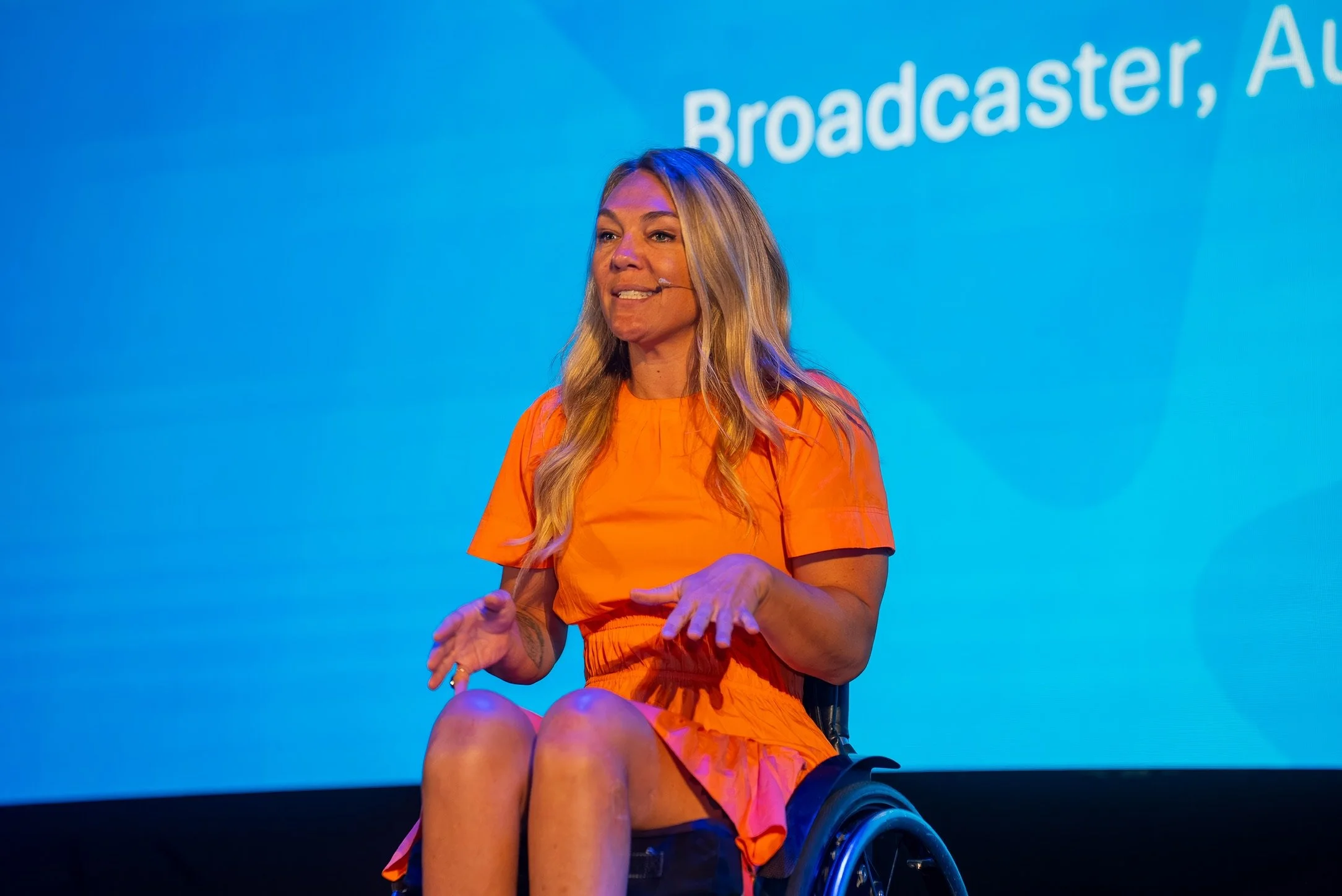 A woman in an orange dress, sitting in a wheelchair, speaking at an event with a blue background that has partial text reading 'Broadcaster, Aut'.