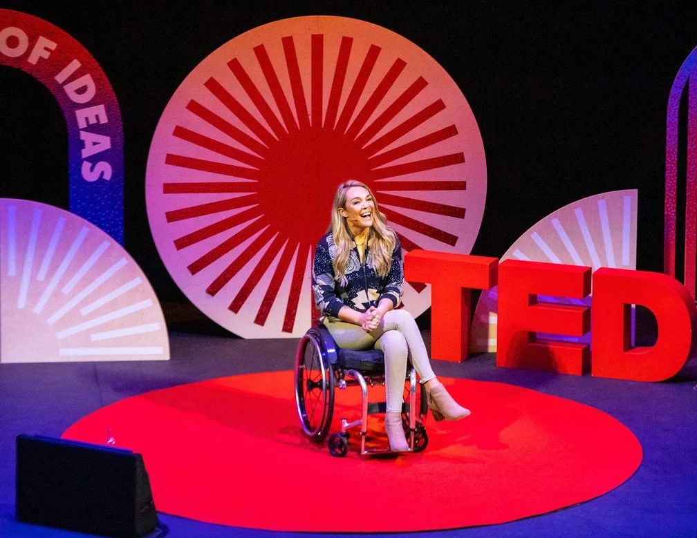 A woman in a wheelchair speaking on a TED stage with a red circular rug. Large red TED letters and colorful circular designs in the background.