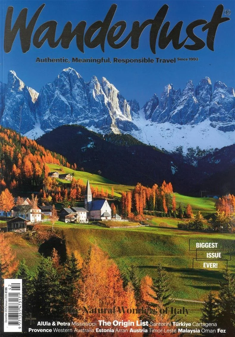 The cover of Wanderlust magazine featuring a scenic mountain landscape with snow-capped peaks, a small church, houses, and autumn trees in a green valley in Italy.
