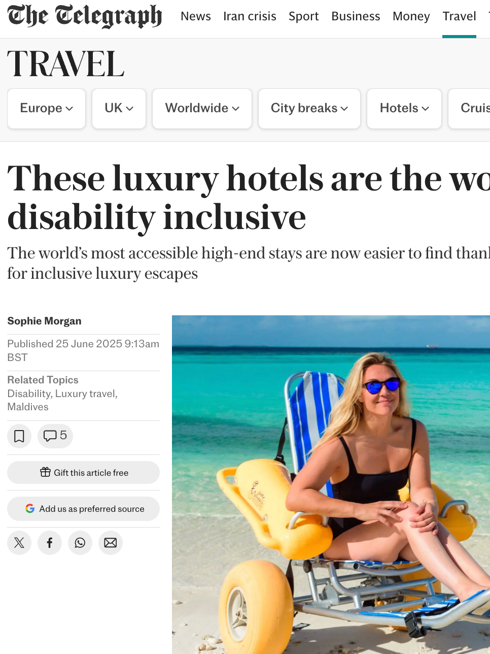 A woman sitting on a beach wheelchair on a sandy beach with turquoise ocean water and a blue sky in the background. She is wearing sunglasses and a black swimsuit.