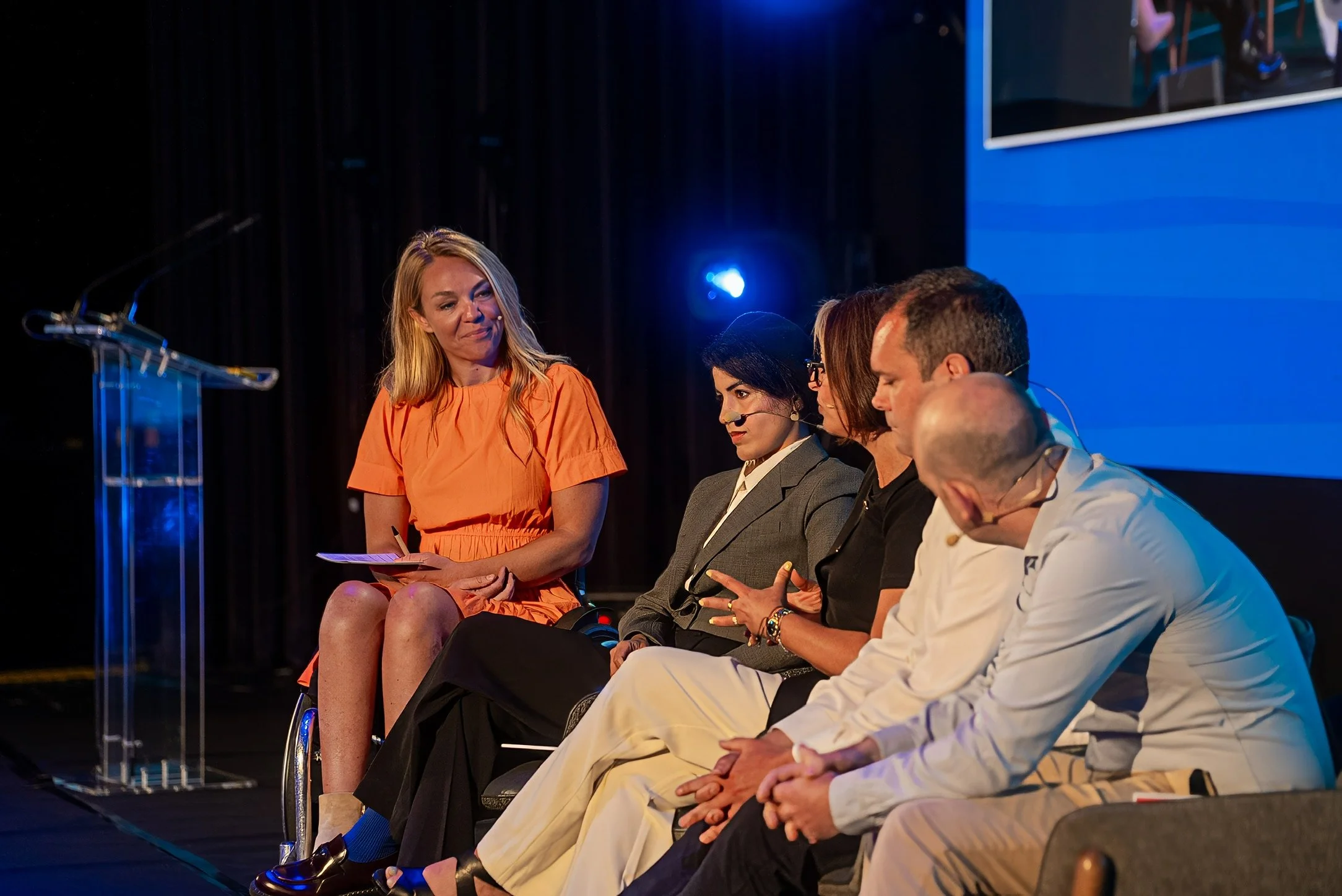 A panel of five people sitting on stage, with one woman in an orange dress holding papers, and the other four individuals listening, with a blue background and a large screen.