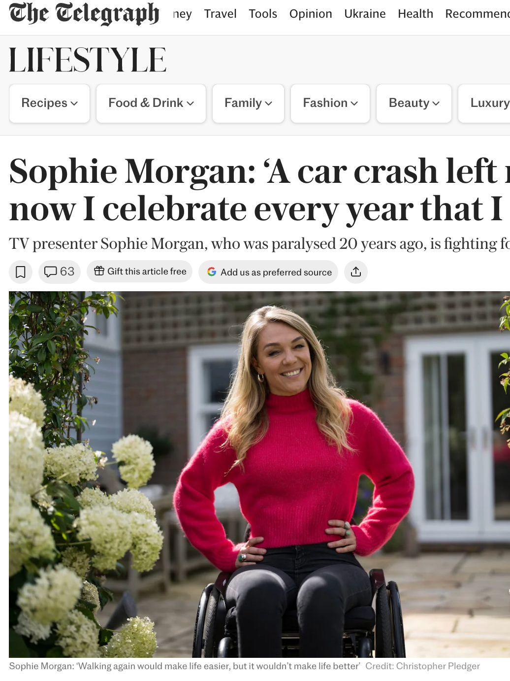 Sophie Morgan, a woman in a pink sweater, sitting in a wheelchair outdoors in a garden with blooming plants and a house in the background, smiling at the camera.