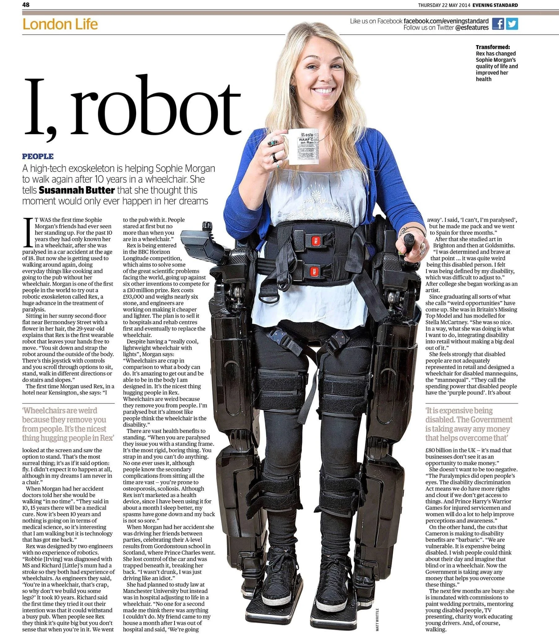 A woman dressed in a blue shirt and black pants, equipped with a robotic exoskeleton for mobility assistance, standing indoors and smiling while holding a cup of coffee.