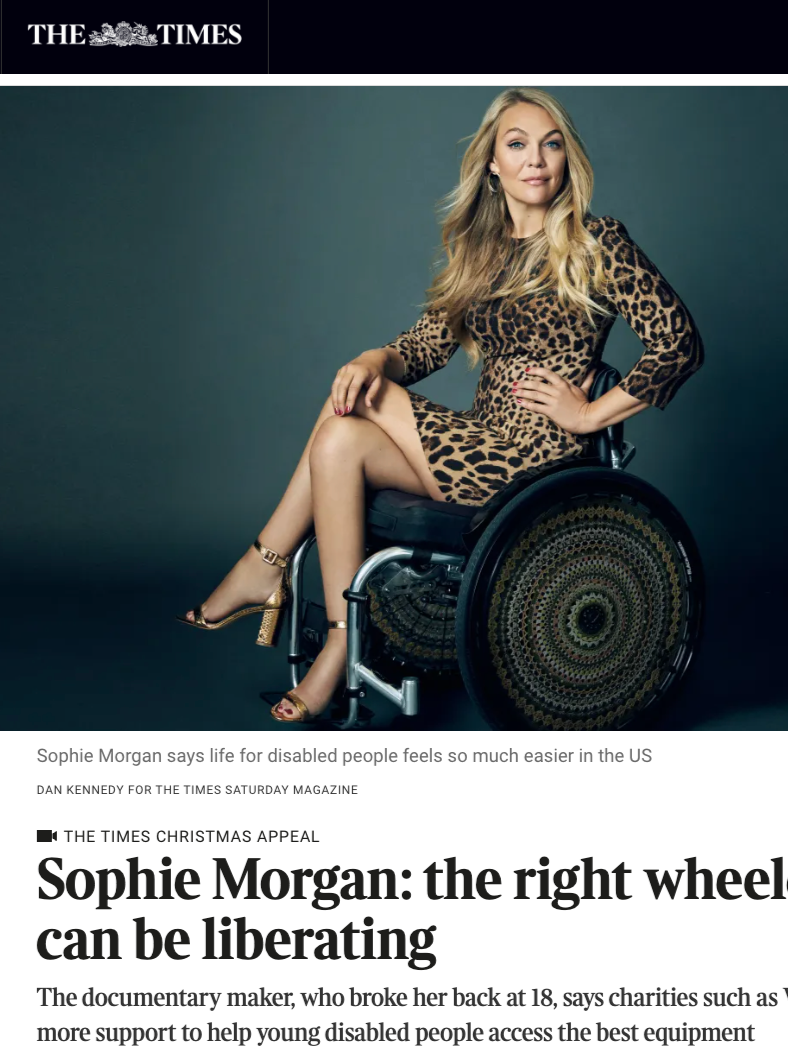 A woman with long blonde hair in a leopard print dress and gold high heels sitting in a wheelchair against a dark background.