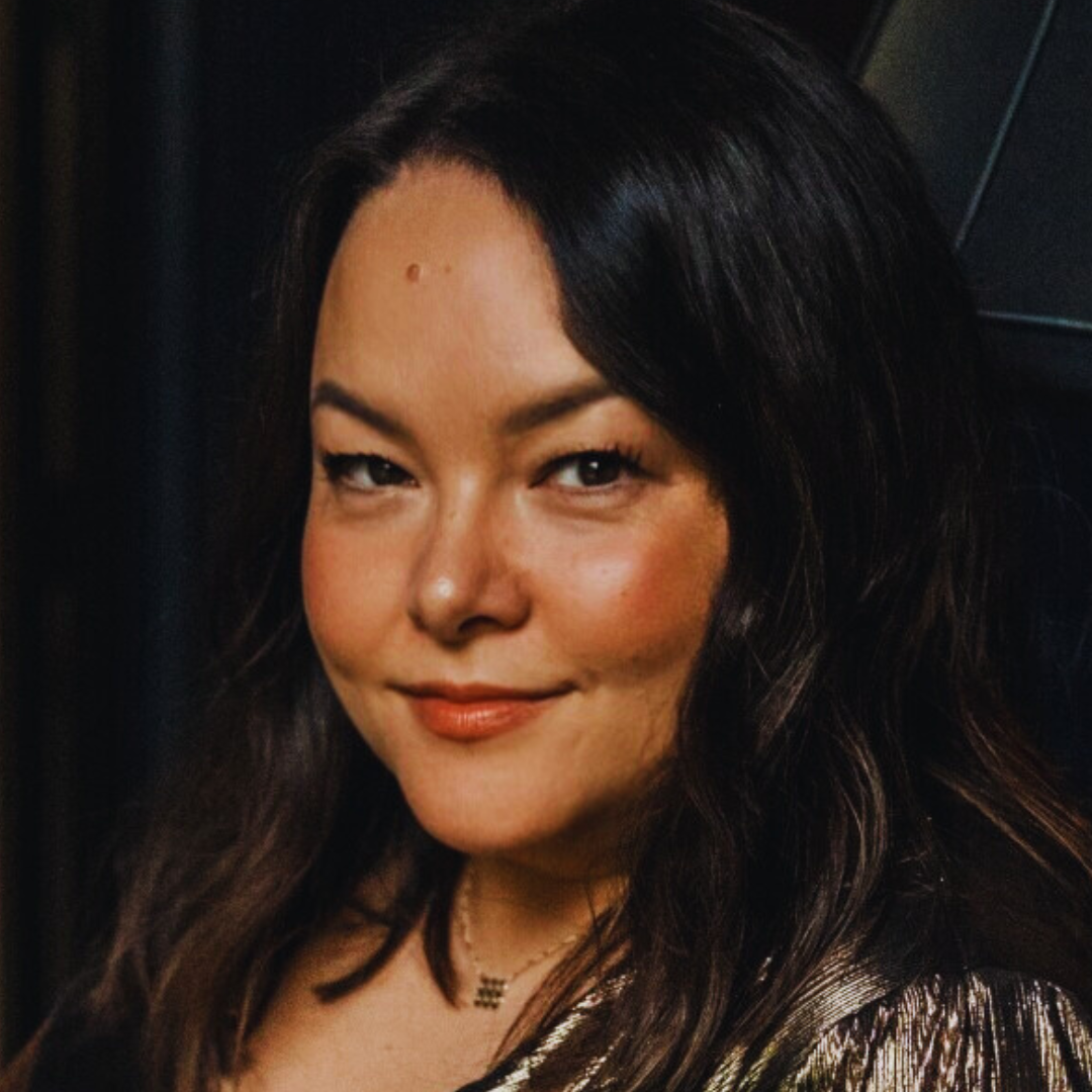Close-up of a woman with dark brown hair, smiling softly, wearing makeup and a shiny top, against a dark background.