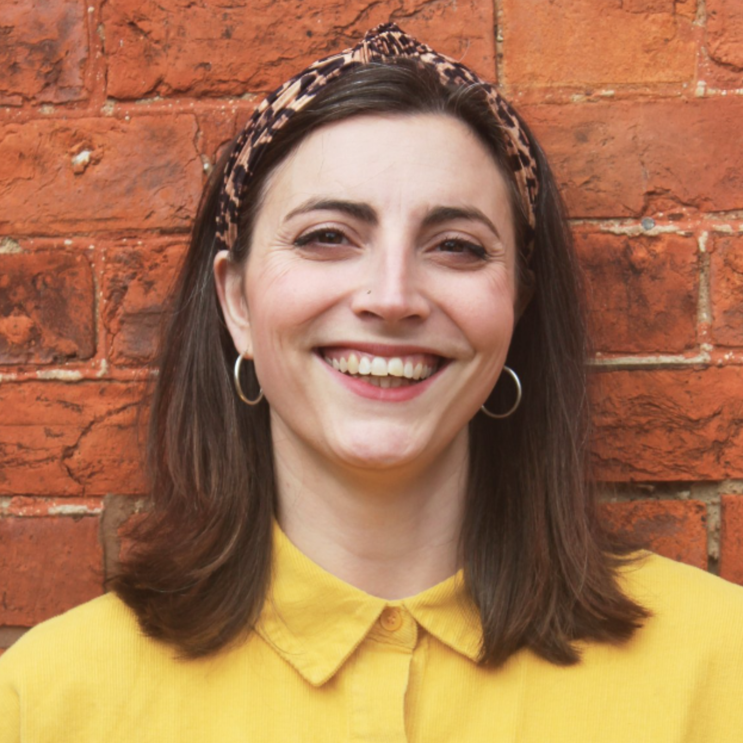 Woman smiling with a headband, wearing a yellow shirt, standing against a brick wall.