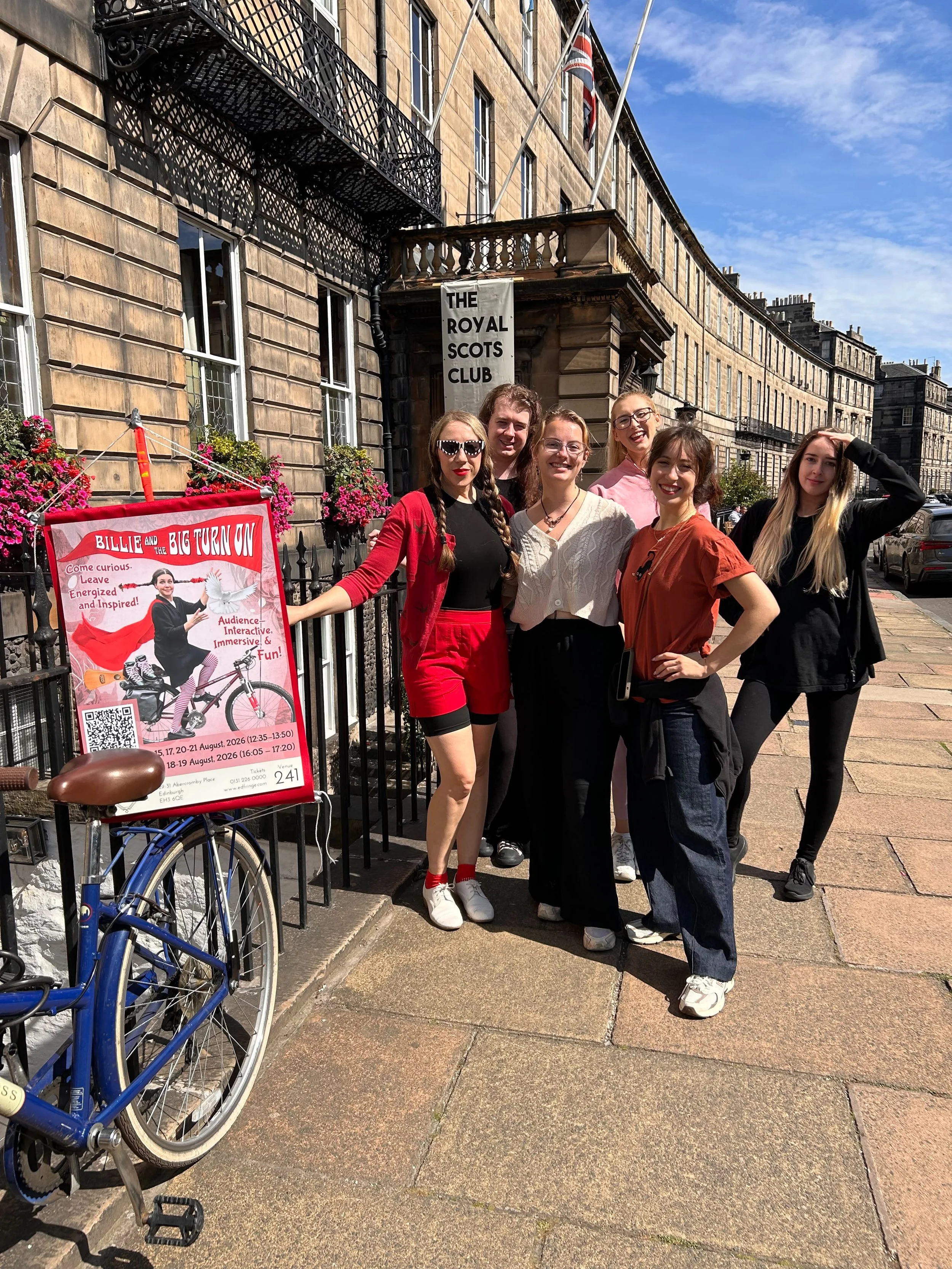 A group of six young women standing outside a historic building with a sign for The Royal Scots Club. They are smiling and posing for the photo. The building has a curved facade, several windows, and flags on the upper balcony. There is a red poster advertising a Bobbie and the Big Turn On event, and a blue bicycle is partially visible in the foreground. The weather is sunny with a partly cloudy sky.