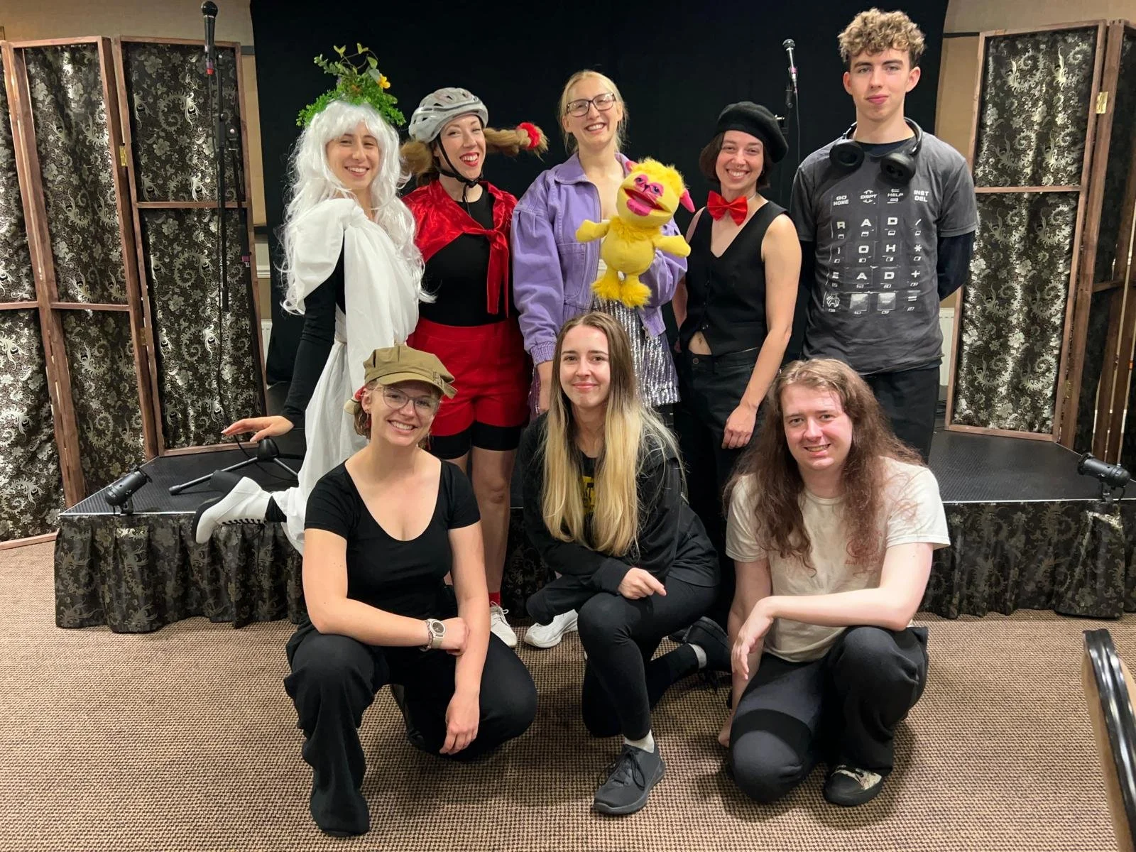 Group of nine young people, some in costumes, posing together in a photo studio with a black backdrop and patterned folding screens.
