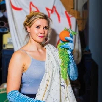 Woman holding a bunch of green grapes with a small orange pumpkin on top, wearing a blue sleeveless top and gloves