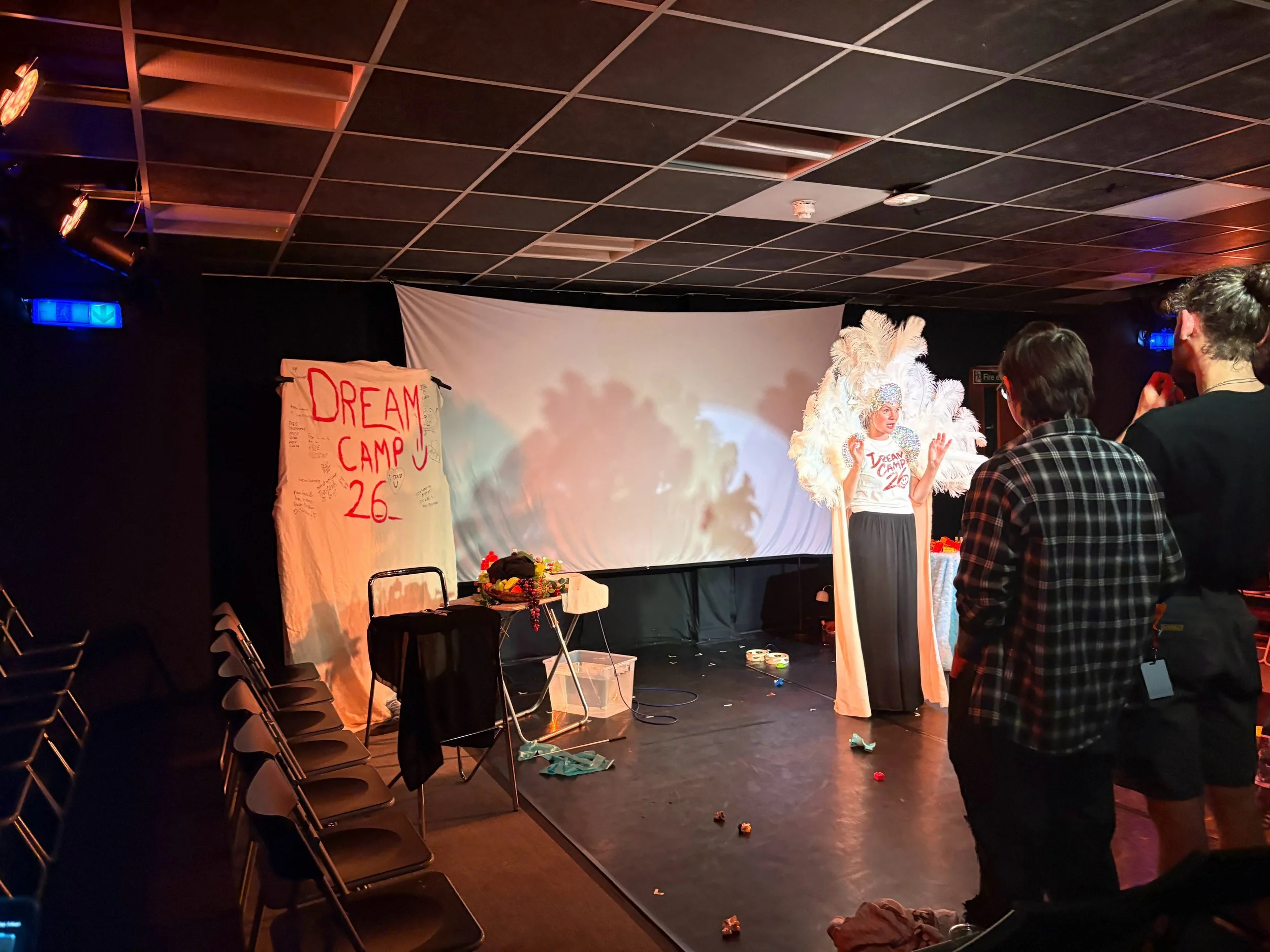 Stage with a white screen, a person dressed in a costume with large feathers, and a sign that reads 'Dream Camp 26.' Several chairs are arranged on the stage, and there are some scattered toys or objects on the floor. A few people are standing and observing.