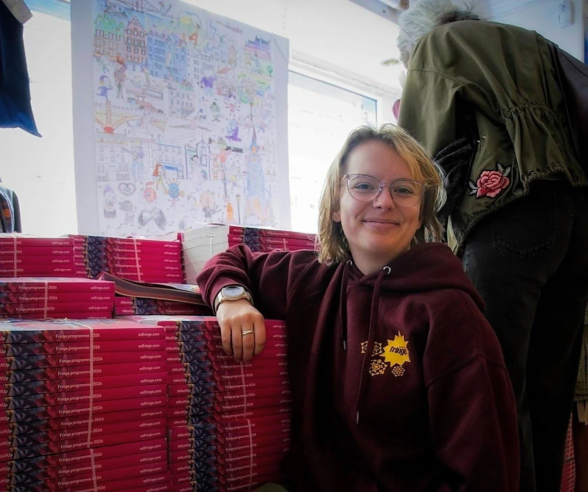 A young woman with glasses and a maroon hoodie smiling at the camera, leaning on a stack of pink boxes labeled 'Fringe program 2026.' Behind her, a man wearing a green jacket with a floral patch is bent over, and a colorful drawing or poster is hanging on the wall near a large window.