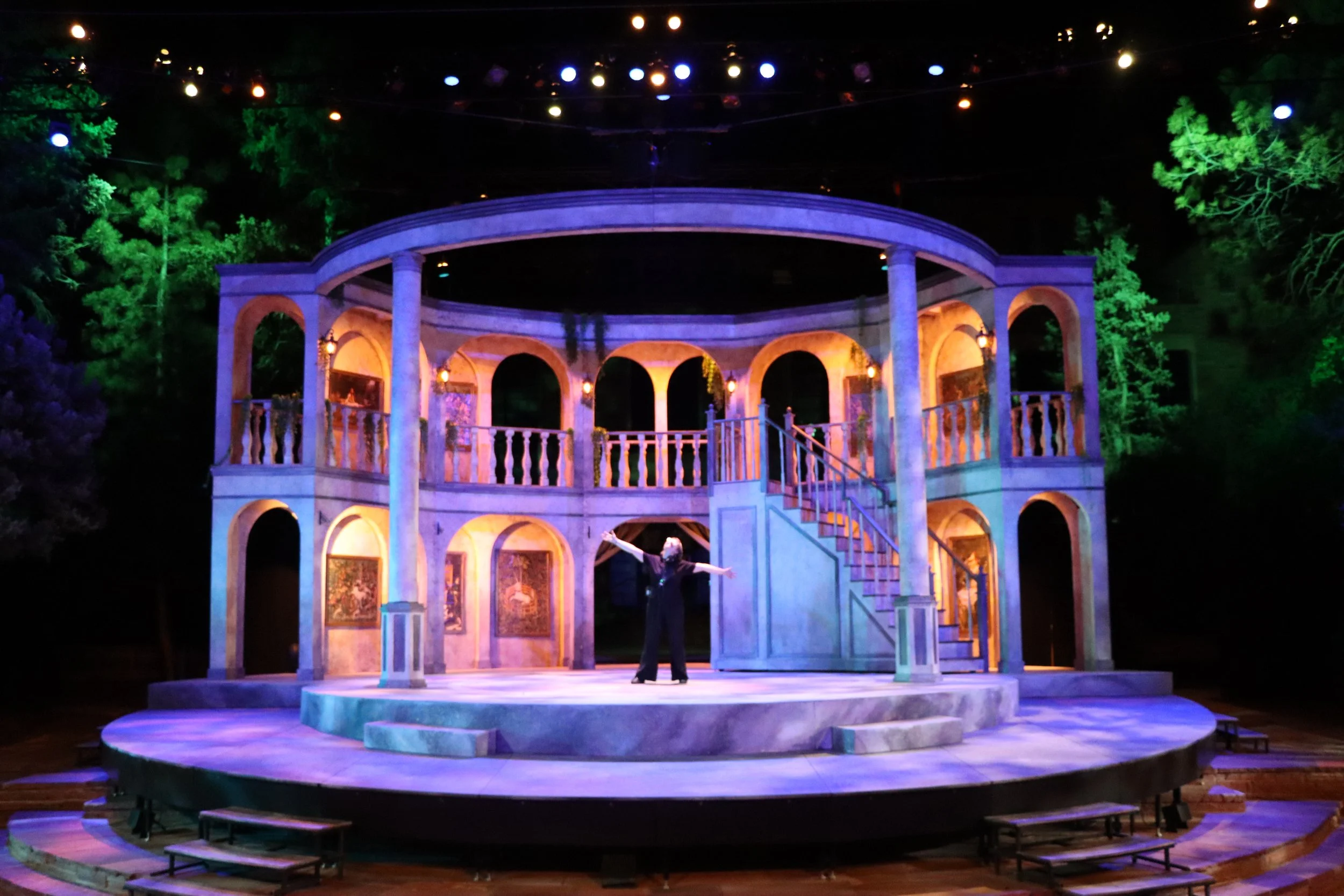 The image shows a stage set themed like a two-story classical house, illuminated with colorful lights, with greenery in the background. A performer is standing in the center with arms outstretched.