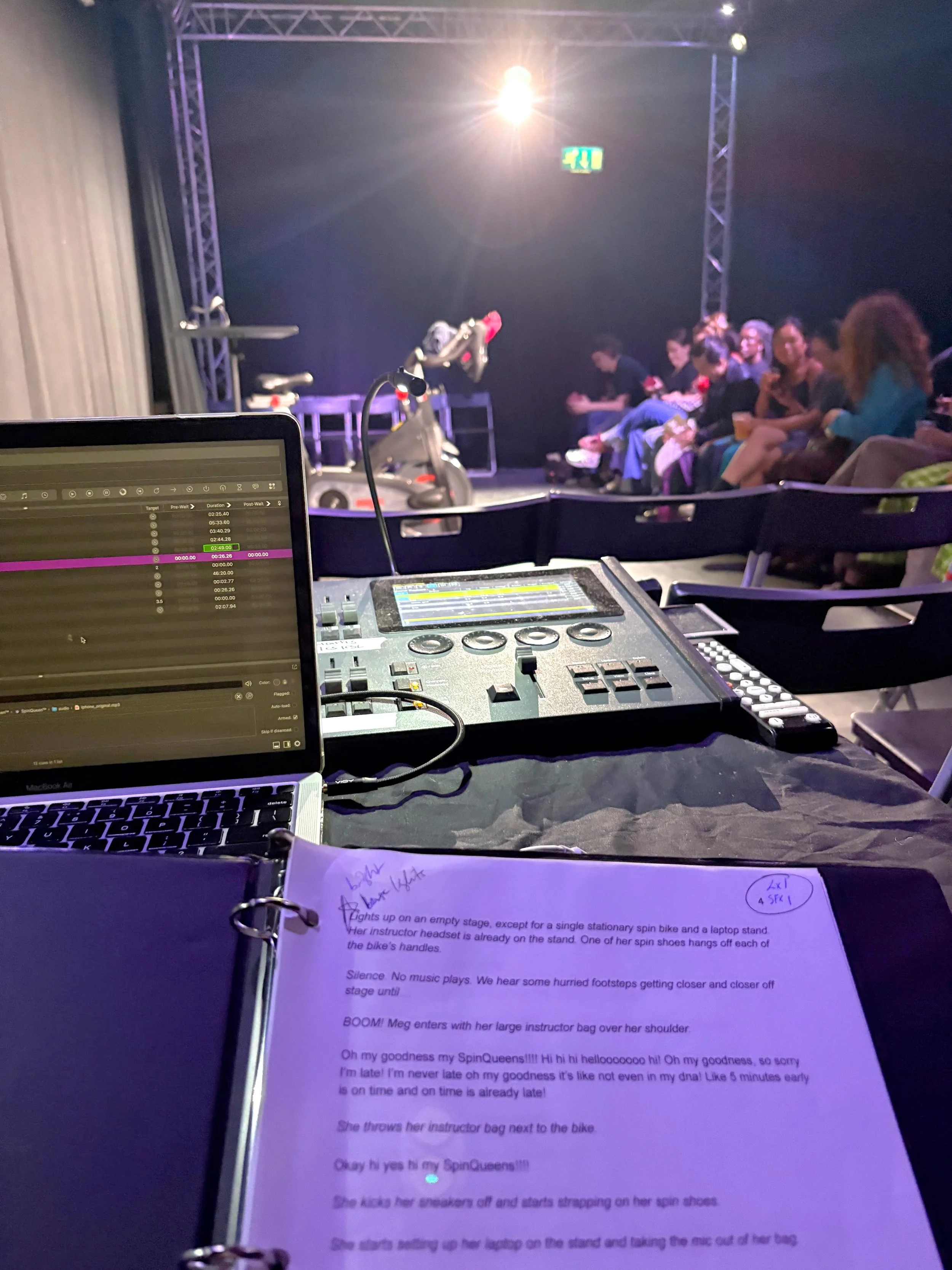 A stage with a music control panel and a laptop in the foreground, and a group of people sitting in the audience in the background. A bright stage light is shining overhead. There are some notes on a paper with typed text partially visible at the bottom.
