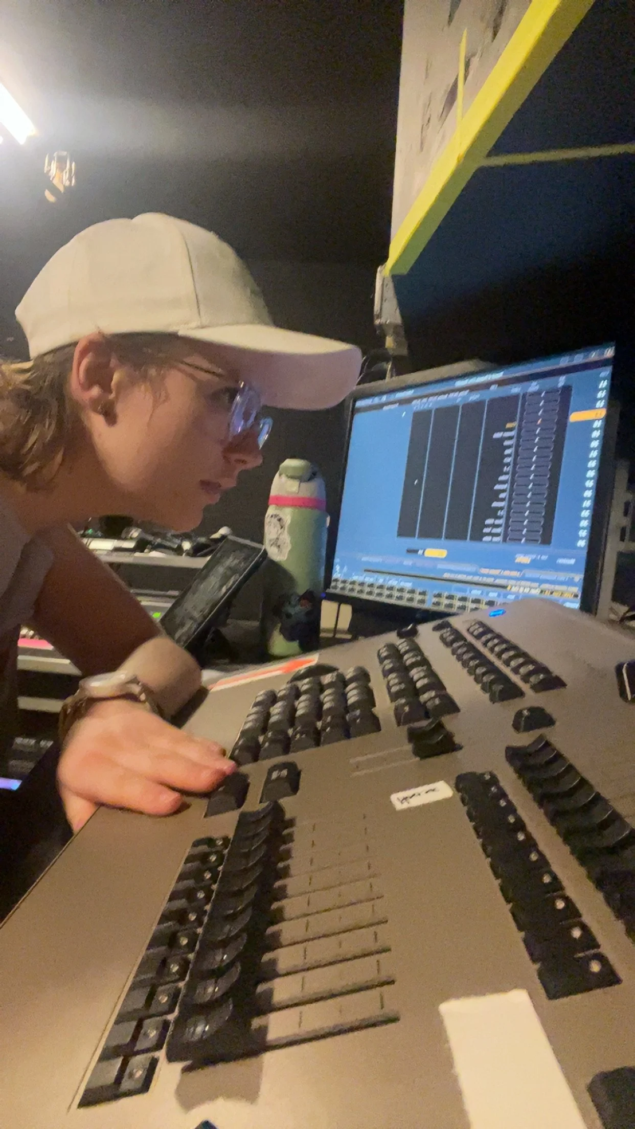 A person working in a music studio, wearing a tan cap, glasses, and a bracelet, using a music production keyboard and working on a computer with digital audio workstation software displayed.