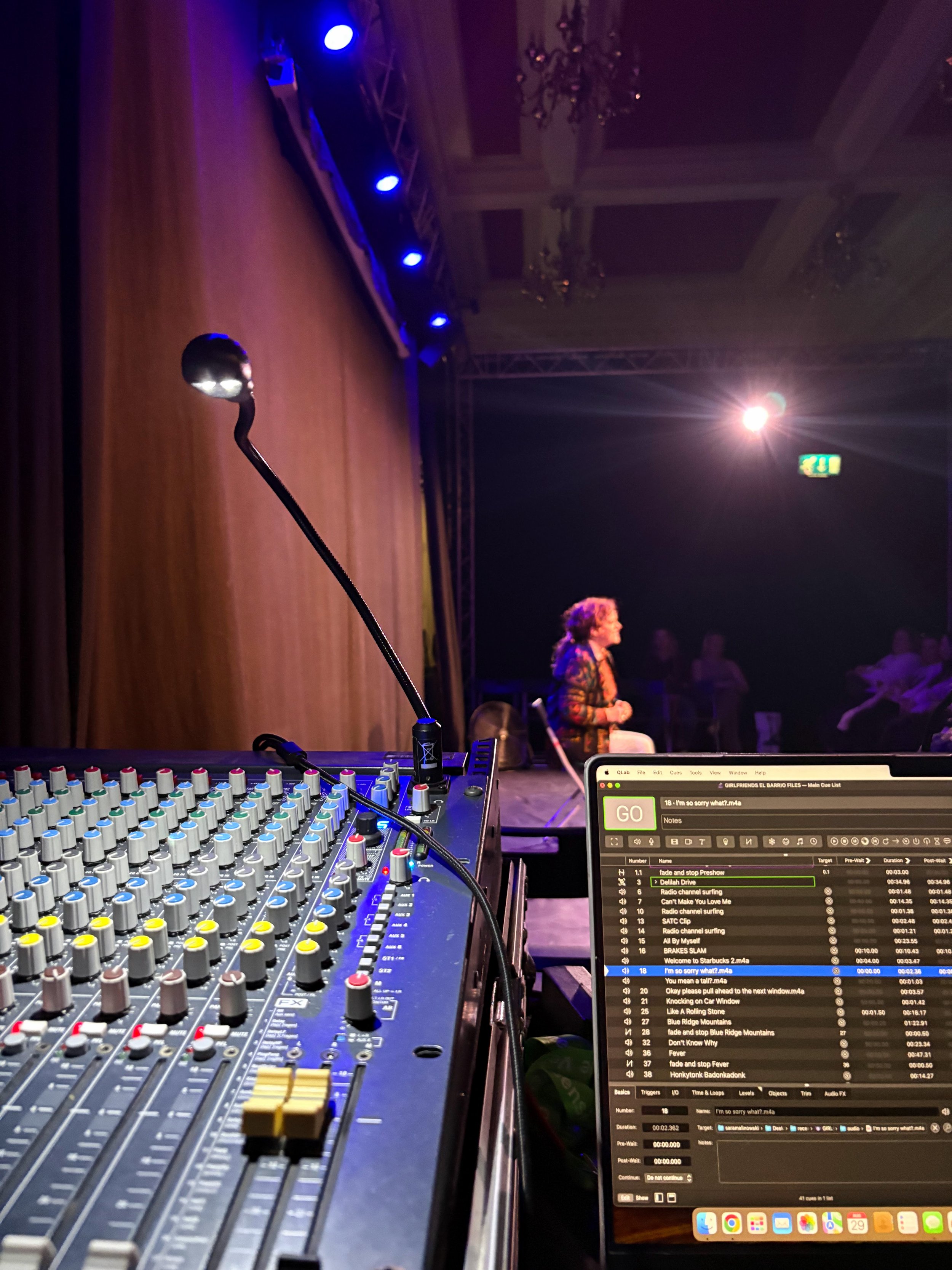 A DJ setup on stage with a sound mixer and a laptop displaying a playlist, with a person in the background under stage lights and chandeliers in a ballroom or event hall.