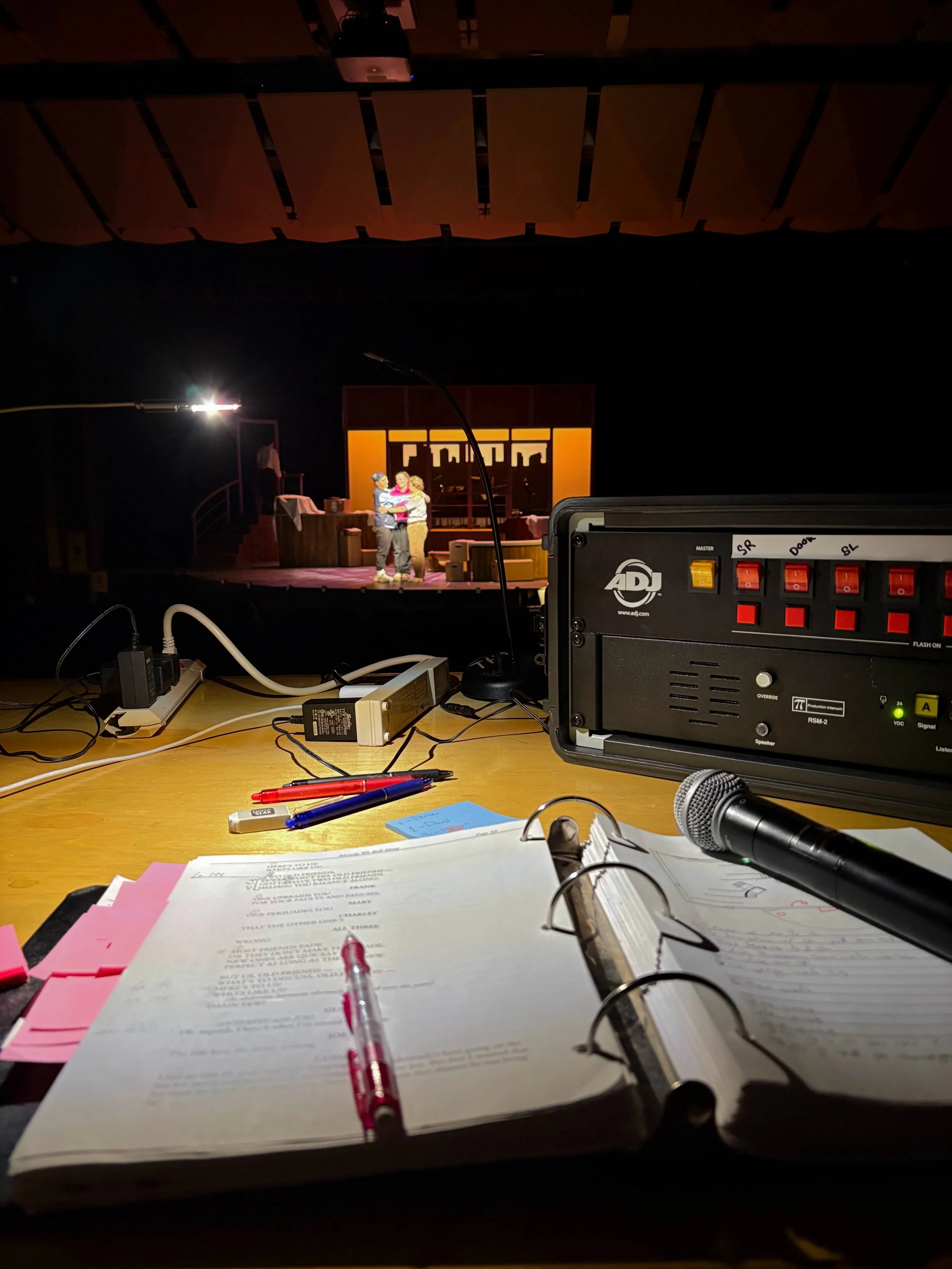 The image shows a view from behind a desk or table with various papers, pens, and a microphone in the foreground. In the background, a stage with three people, possibly actors, standing close together, can be seen. The stage has a set with a window and some furniture.