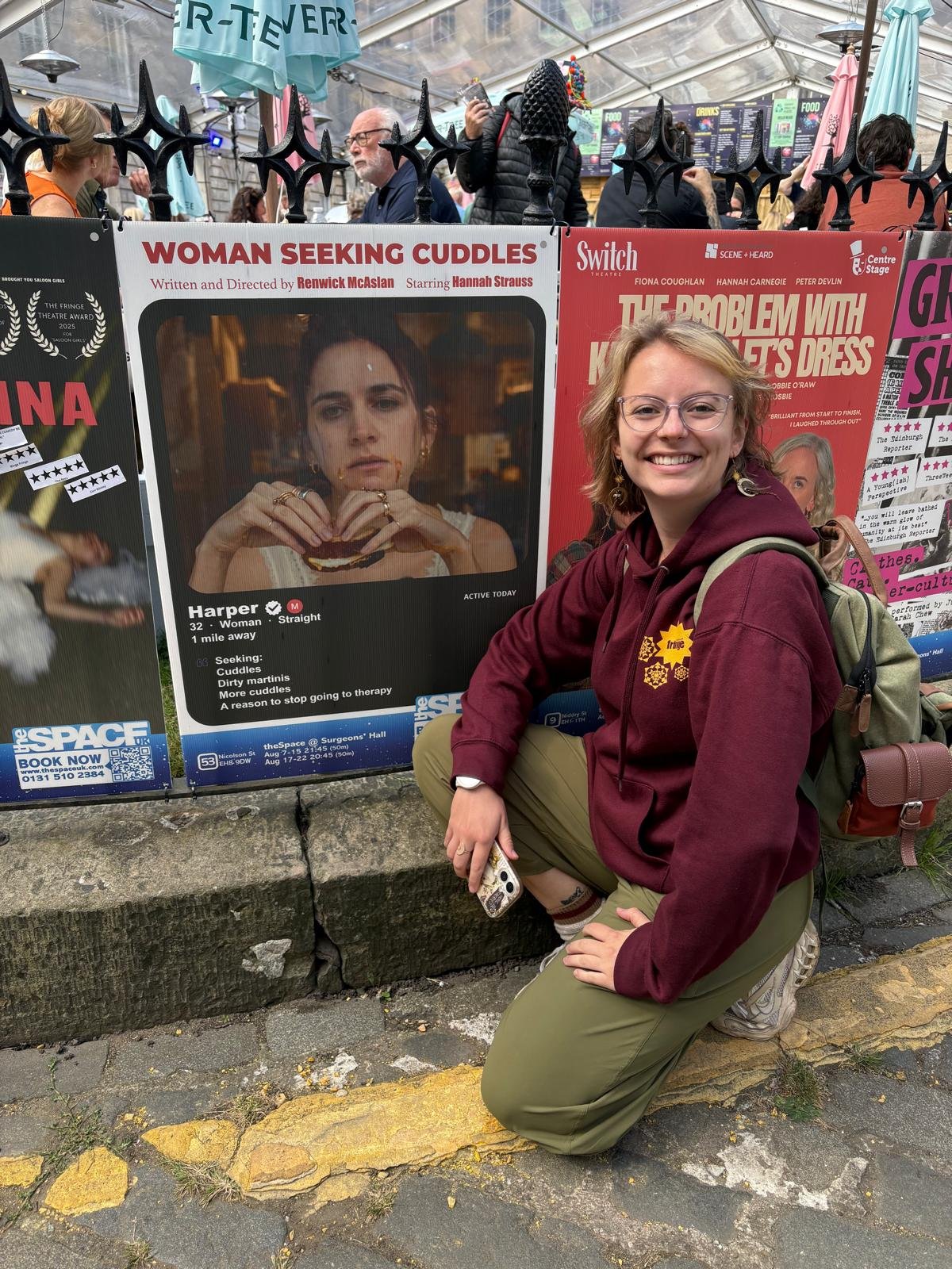A young woman with glasses and short hair, wearing a maroon hoodie and green pants, kneeling on the sidewalk in front of a movie poster for a play titled "Woman Seeking Cuddles." She is smiling and holding a smartphone, with a backpack on her back. The poster features a woman holding a burger, and there are other people and colorful umbrellas visible overhead.
