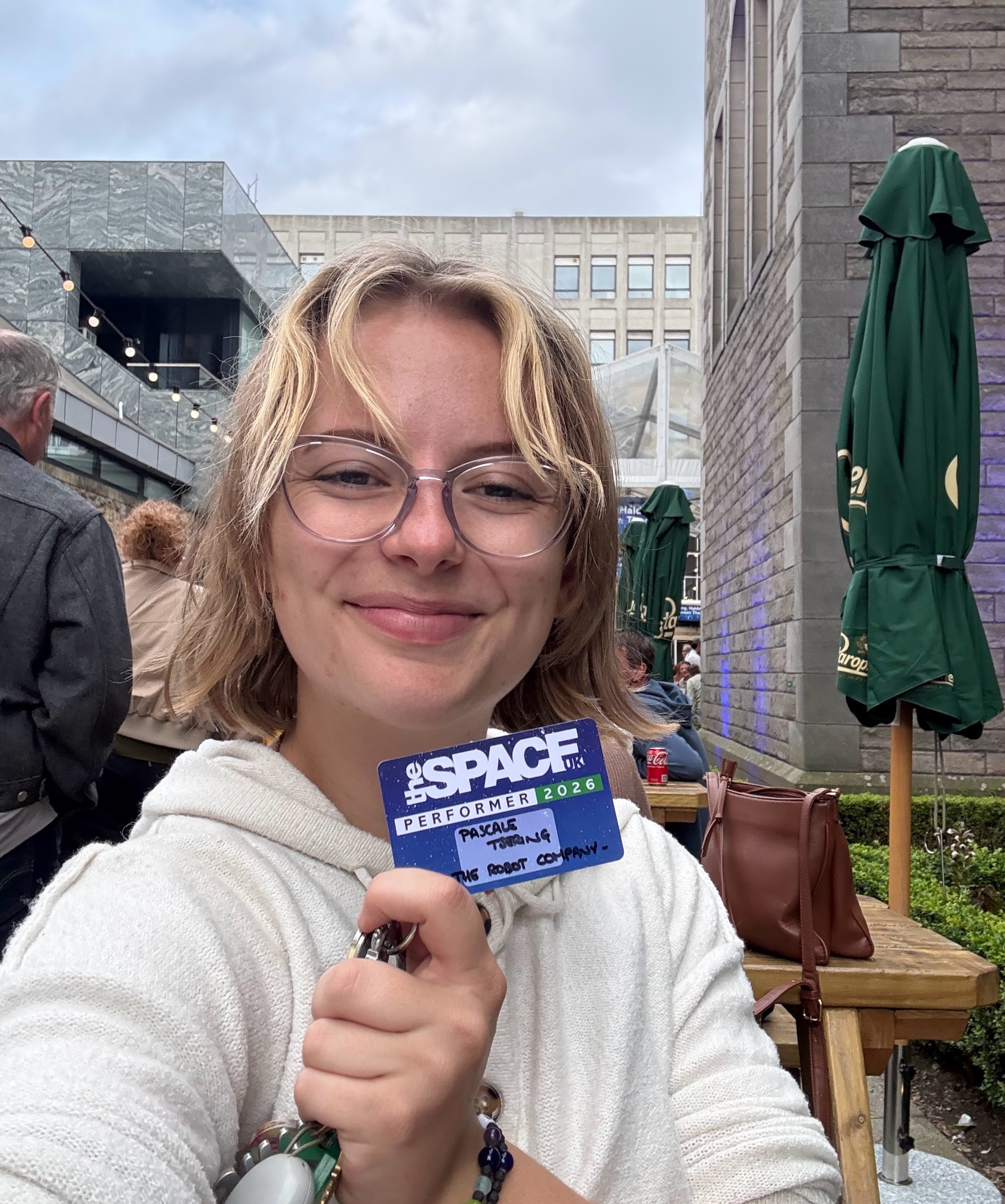 A woman taking a selfie outdoors, smiling, with a crowd behind her. She holds a badge that reads 'the SPACE UK Performer 2026' with handwritten notes about 'Payscale, Tour, The Robot Company.' There are green umbrellas and modern buildings in the background on a cloudy day.