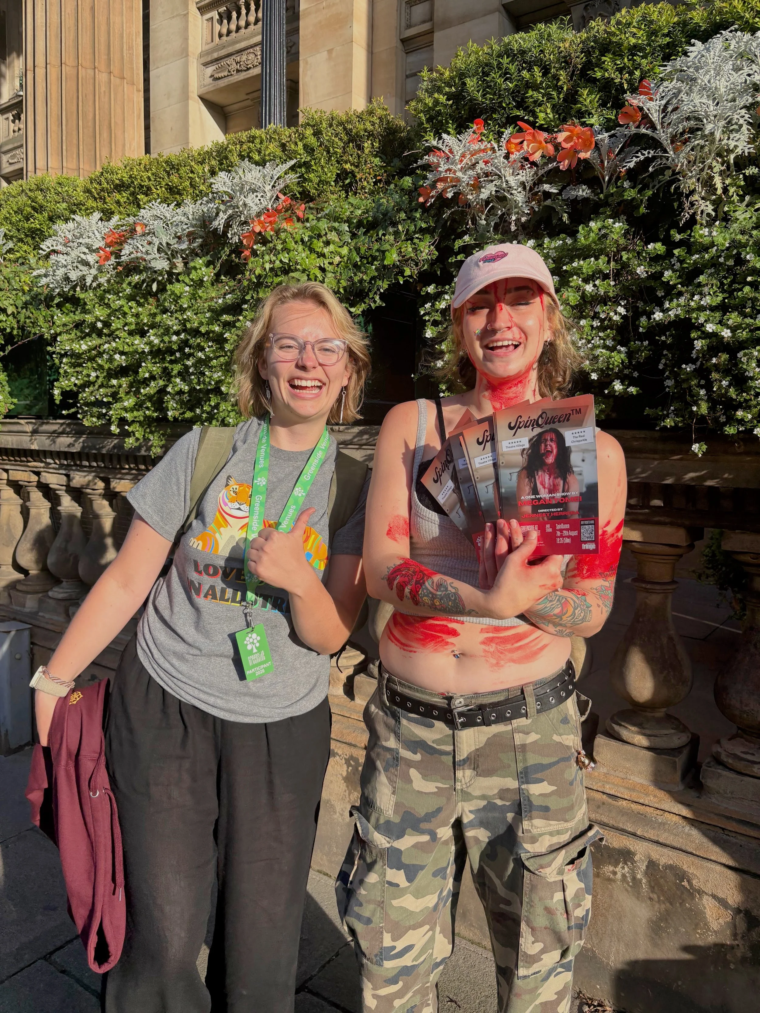 Two young women smiling, standing outdoors in front of a garden hedge with flowers and stone architecture. One is giving a thumbs up, the other is holding flyers, both appear to be at a public event or gathering.