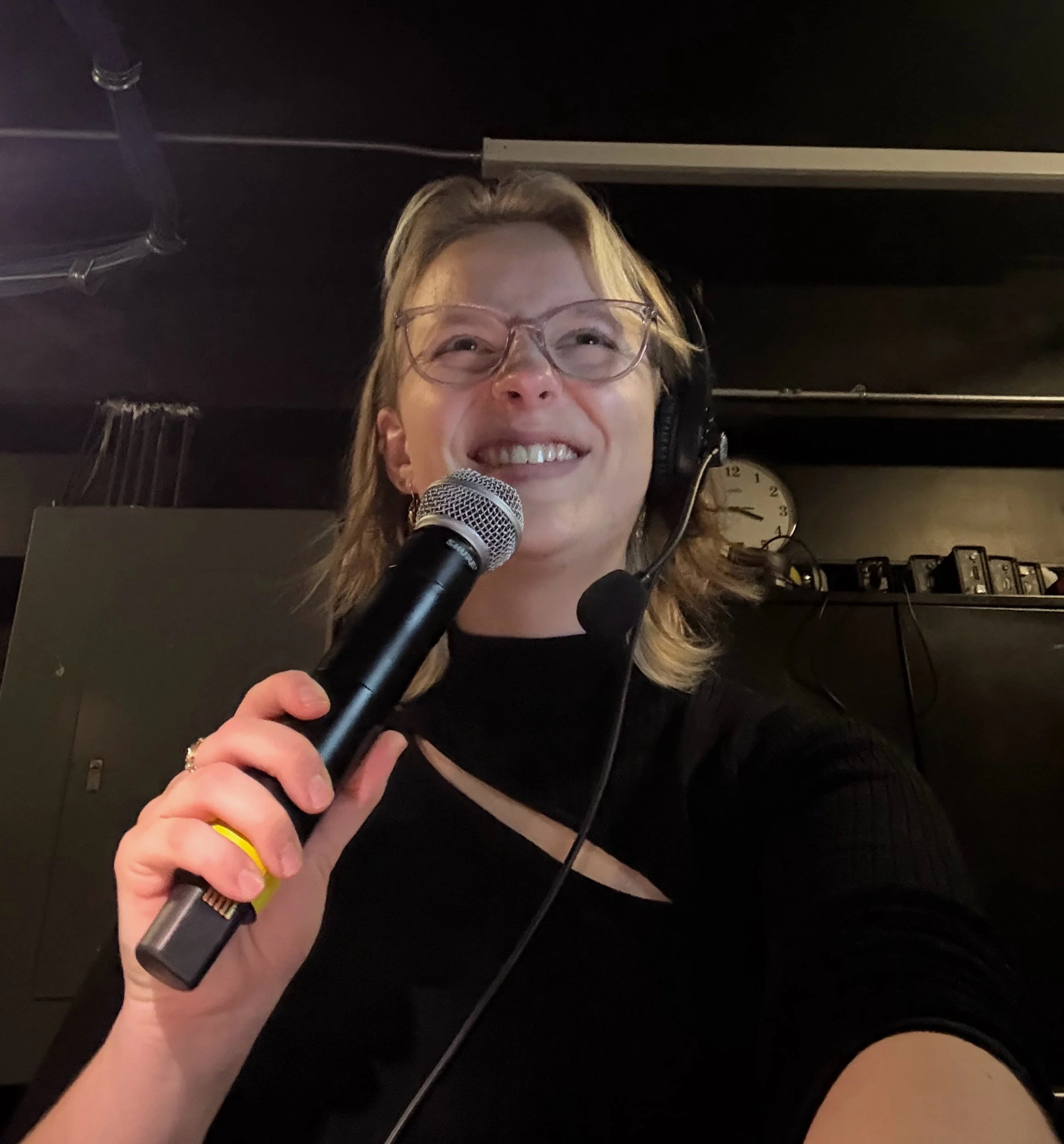 A person holding a microphone with both hands, wearing glasses and headphones, smiling and squinting in a room with a black ceiling and a clock.