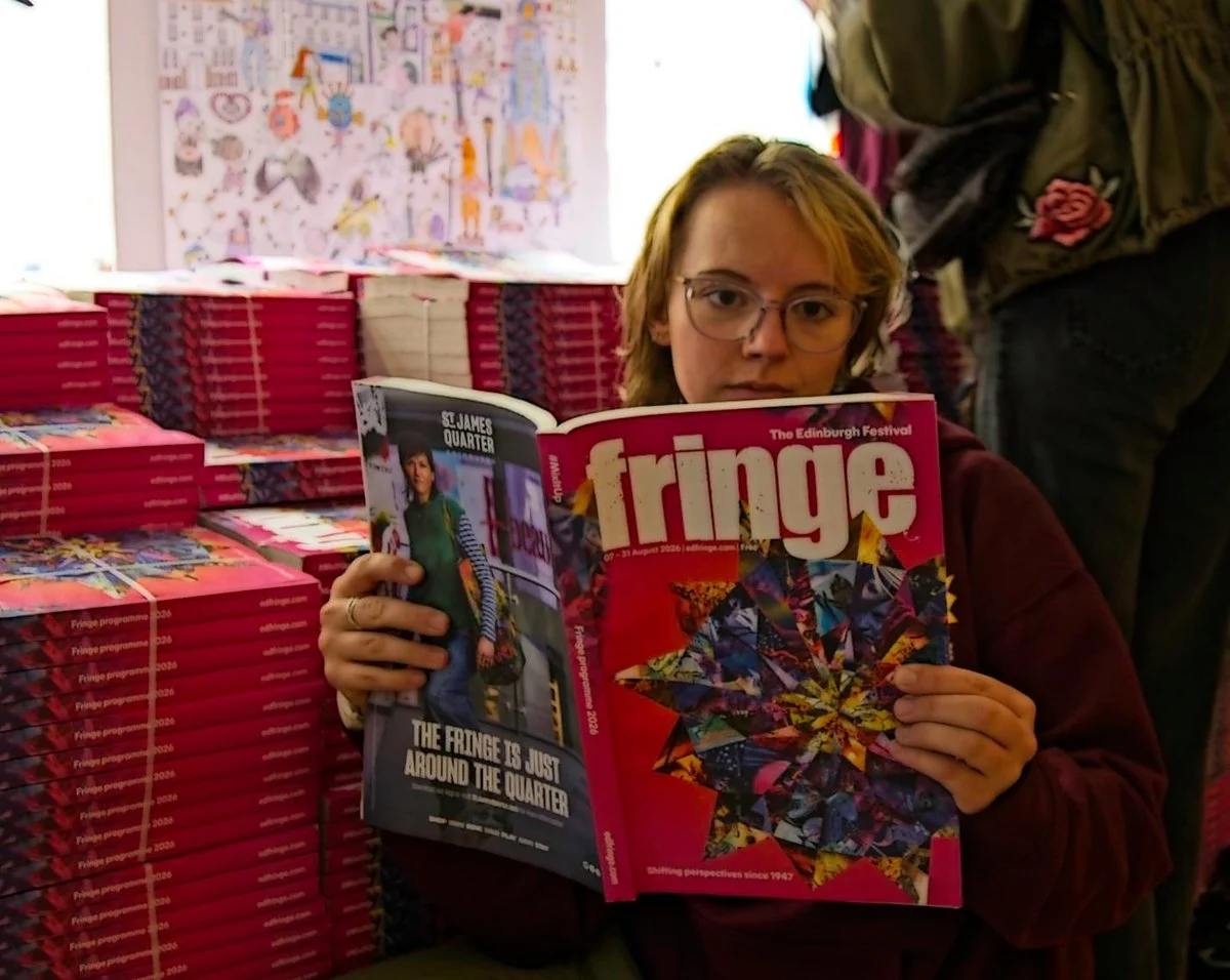 A young woman with glasses and blonde hair holding a copy of Fringe magazine at the Edinburgh Festival, with stacks of the magazine behind her and a colorful poster on the wall.
