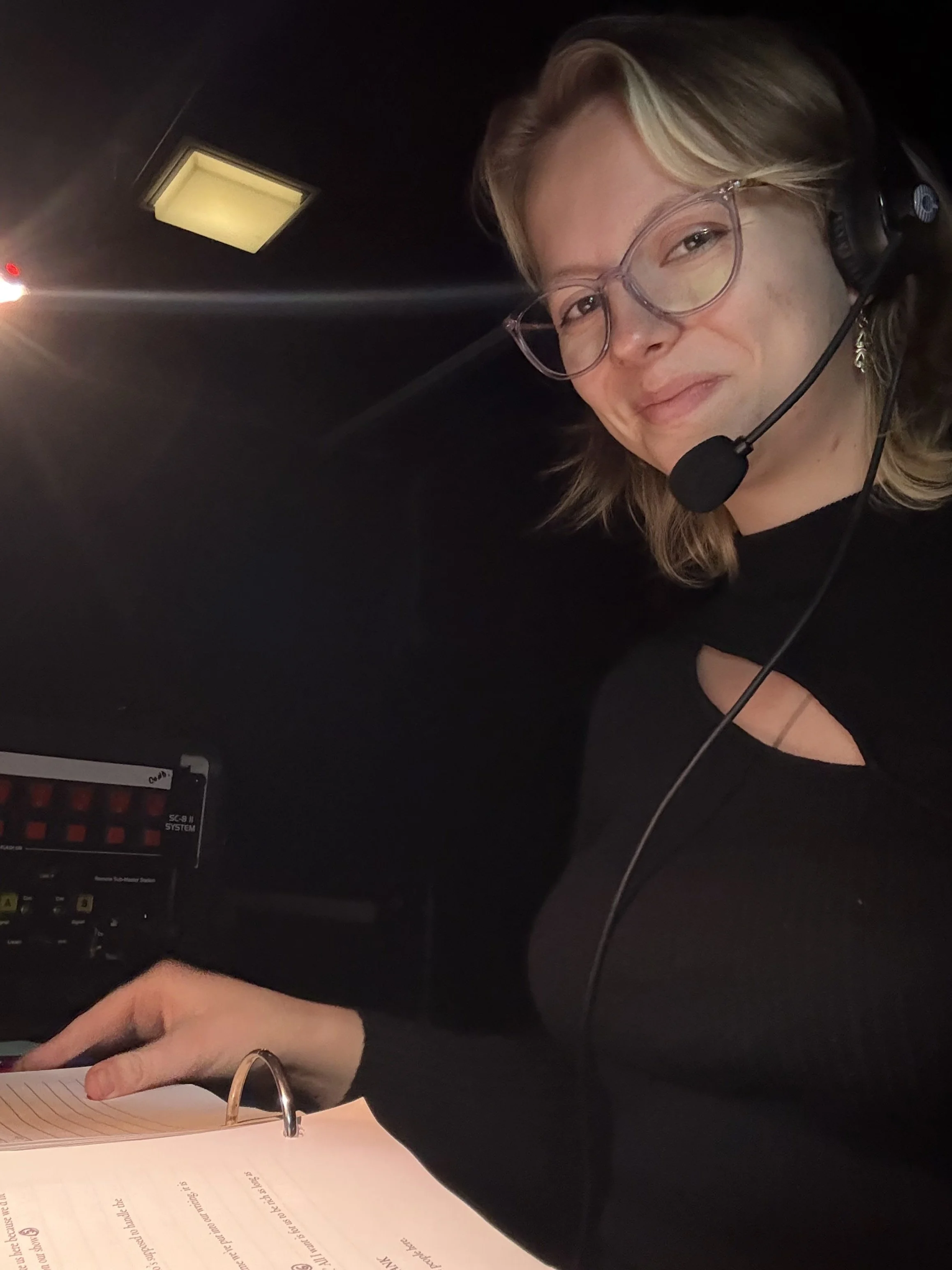 A woman wearing glasses and headphones is smiling while looking at a document or book in front of her. The background is dark and a light is visible on the ceiling.