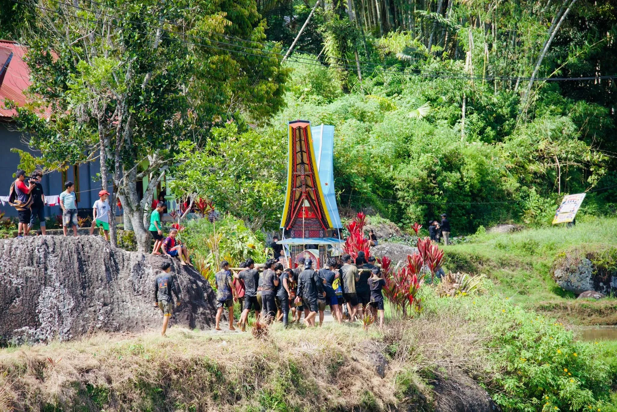Group of people gathered around a traditional Balinese Cremation Tower in lush, tropical greenery, with some standing on rocks and others walking nearby.